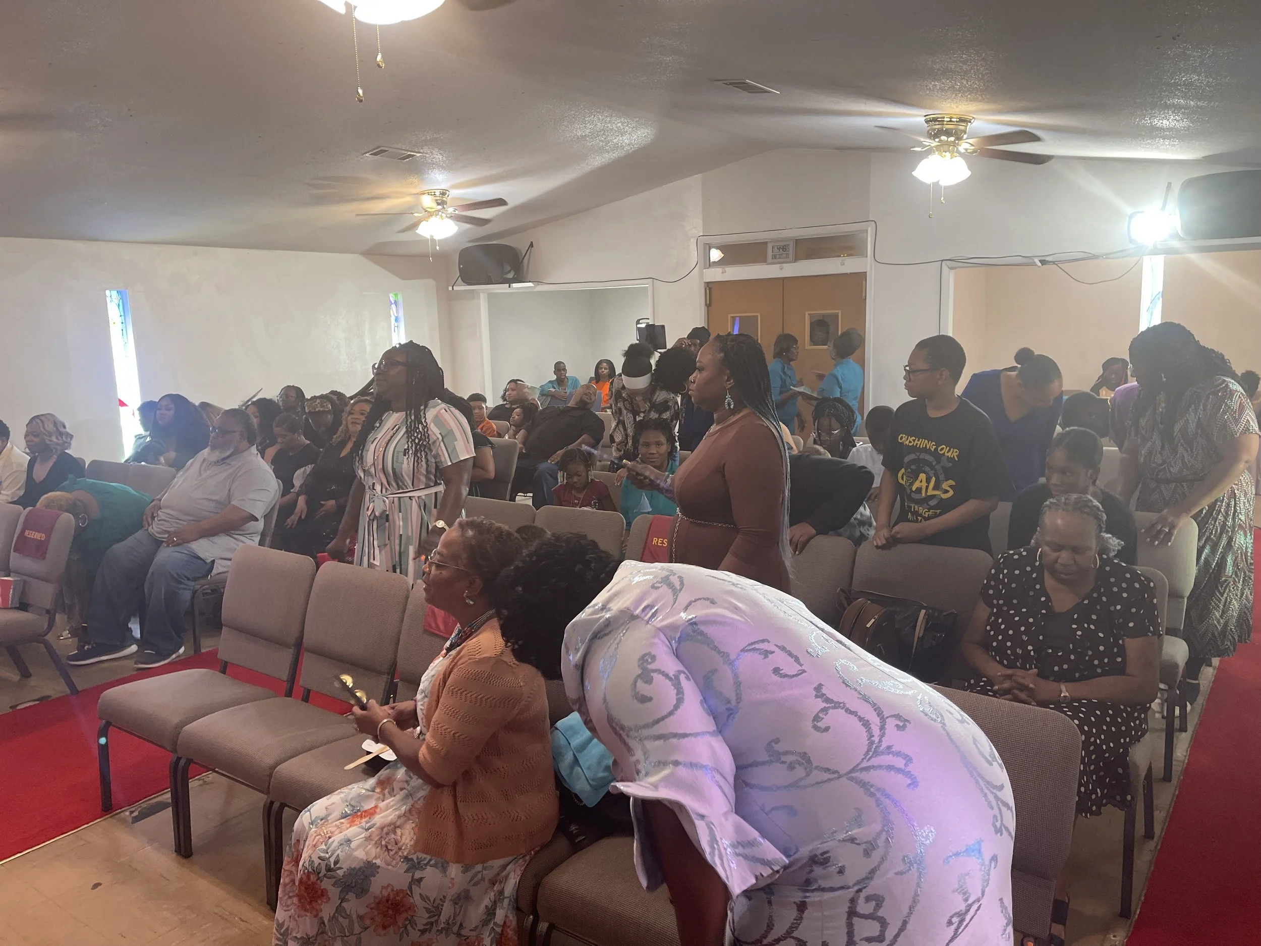A congregation gathered in a church or community hall, with many people standing and sitting, some with heads bowed or engaged in prayer, in a room with ceiling fans and stained glass windows.