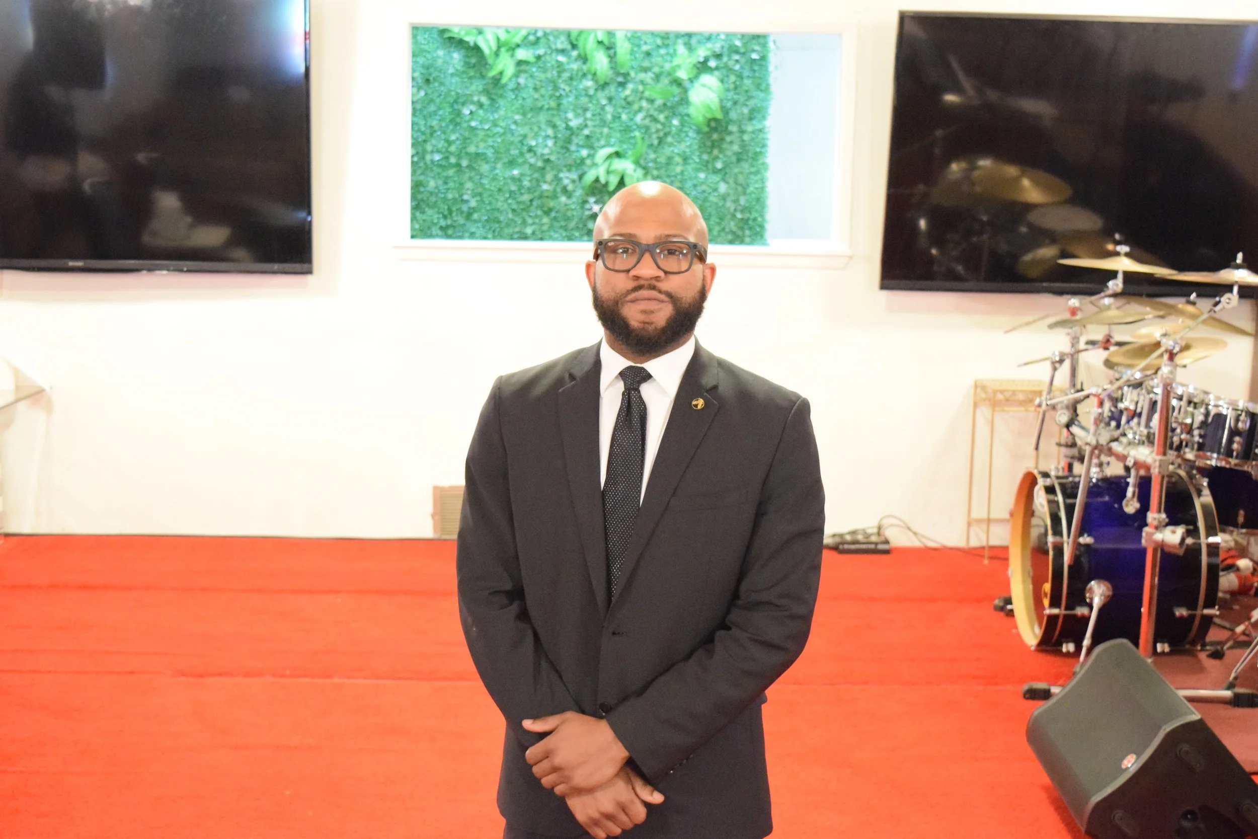 A man in a black suit, white shirt, and black tie standing on a red carpeted stage, wearing glasses and sporting a beard. Behind him are two large mounted flat-screen TVs, a window with a green landscape outside, and a drum set with cymbals and drums.