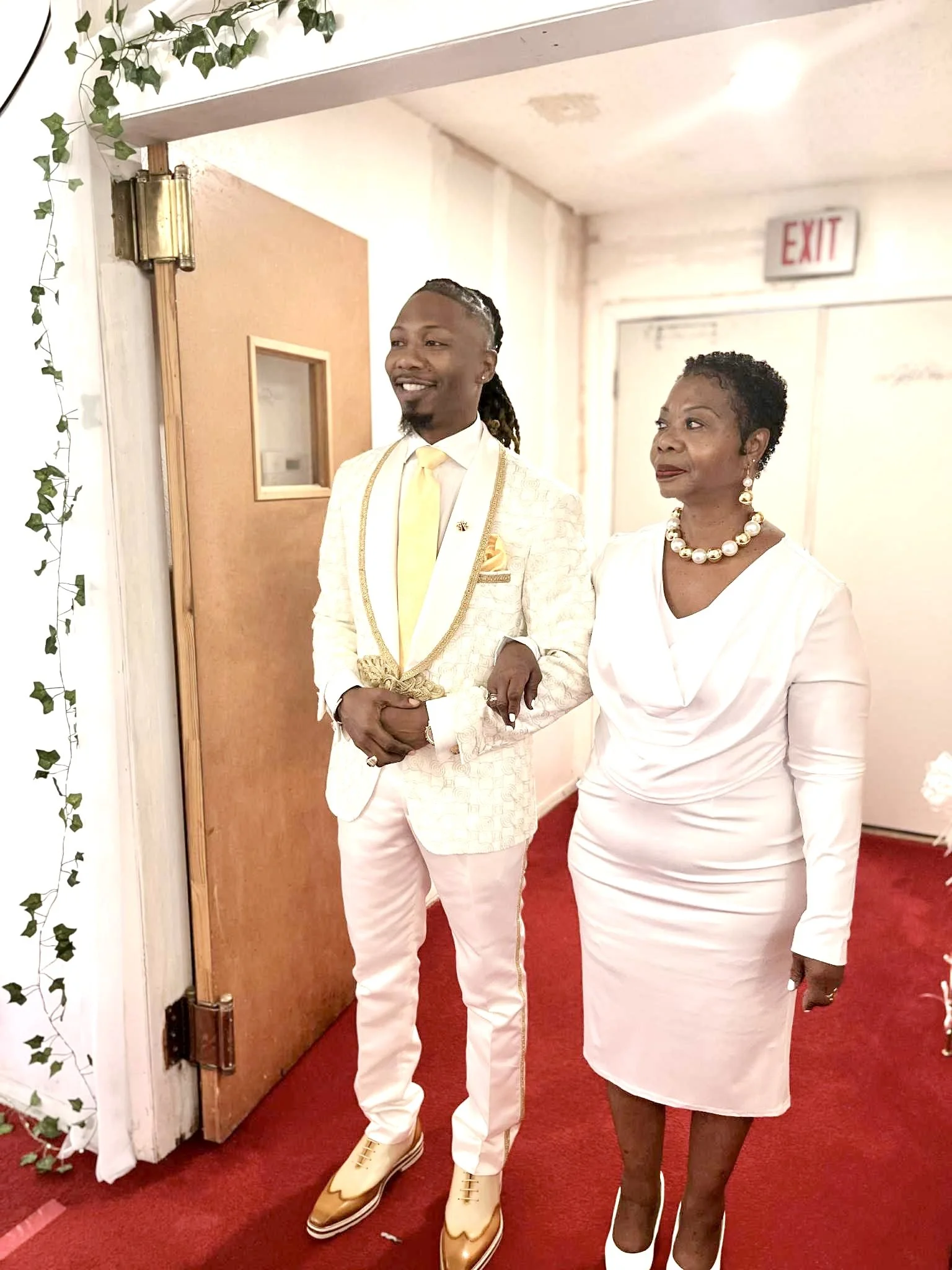 A young man in a white suit with gold accents, yellow tie, and gold shoes standing with a woman in a white dress, pearl necklace, and earrings. They are indoors near a door with an exit sign above.