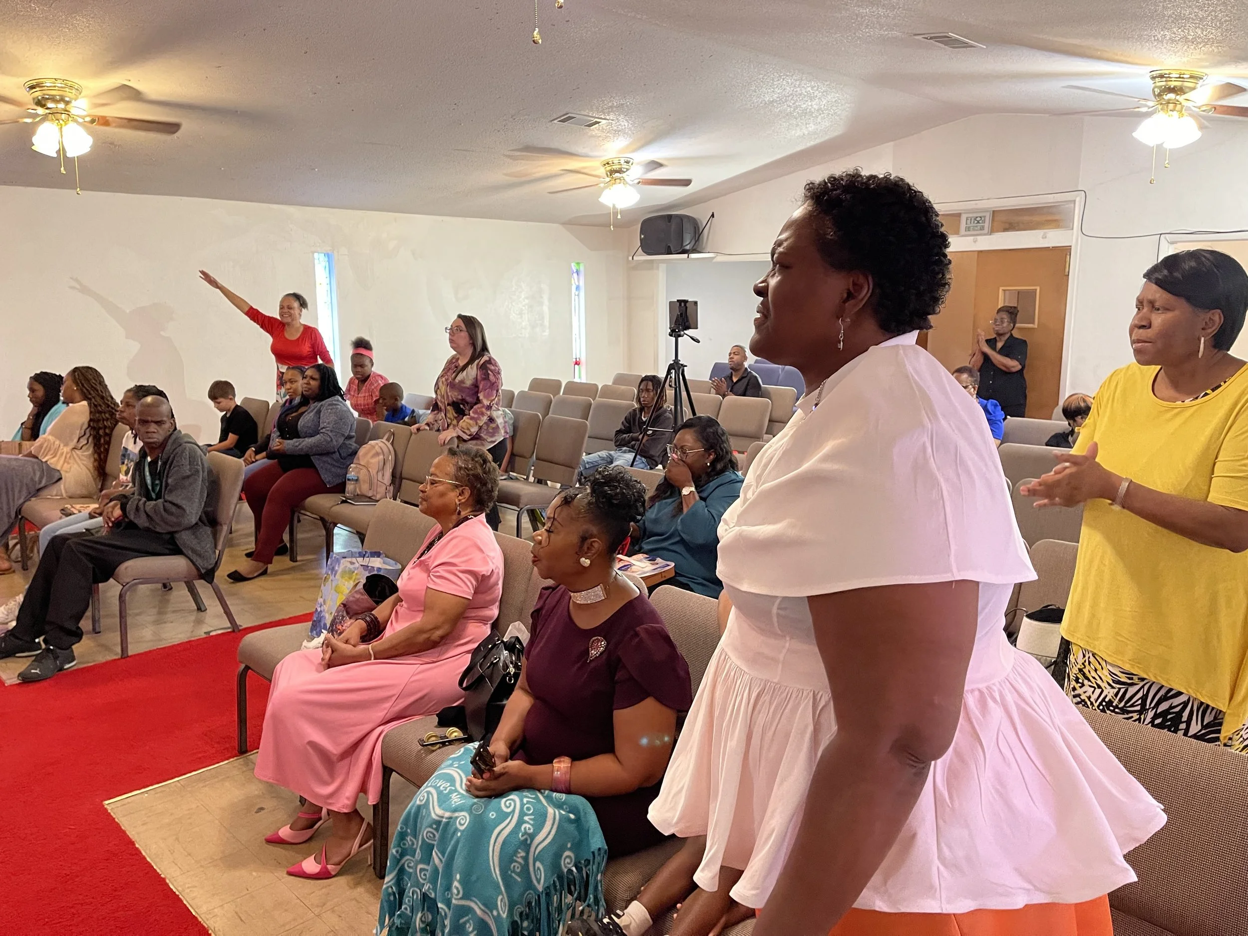 People gathered in a church or community hall, some standing and some seated, during a service or event. The room has ceiling fans, windows, and chairs arranged on a red carpet. Several women and children are present, some engaged in prayer or singing.