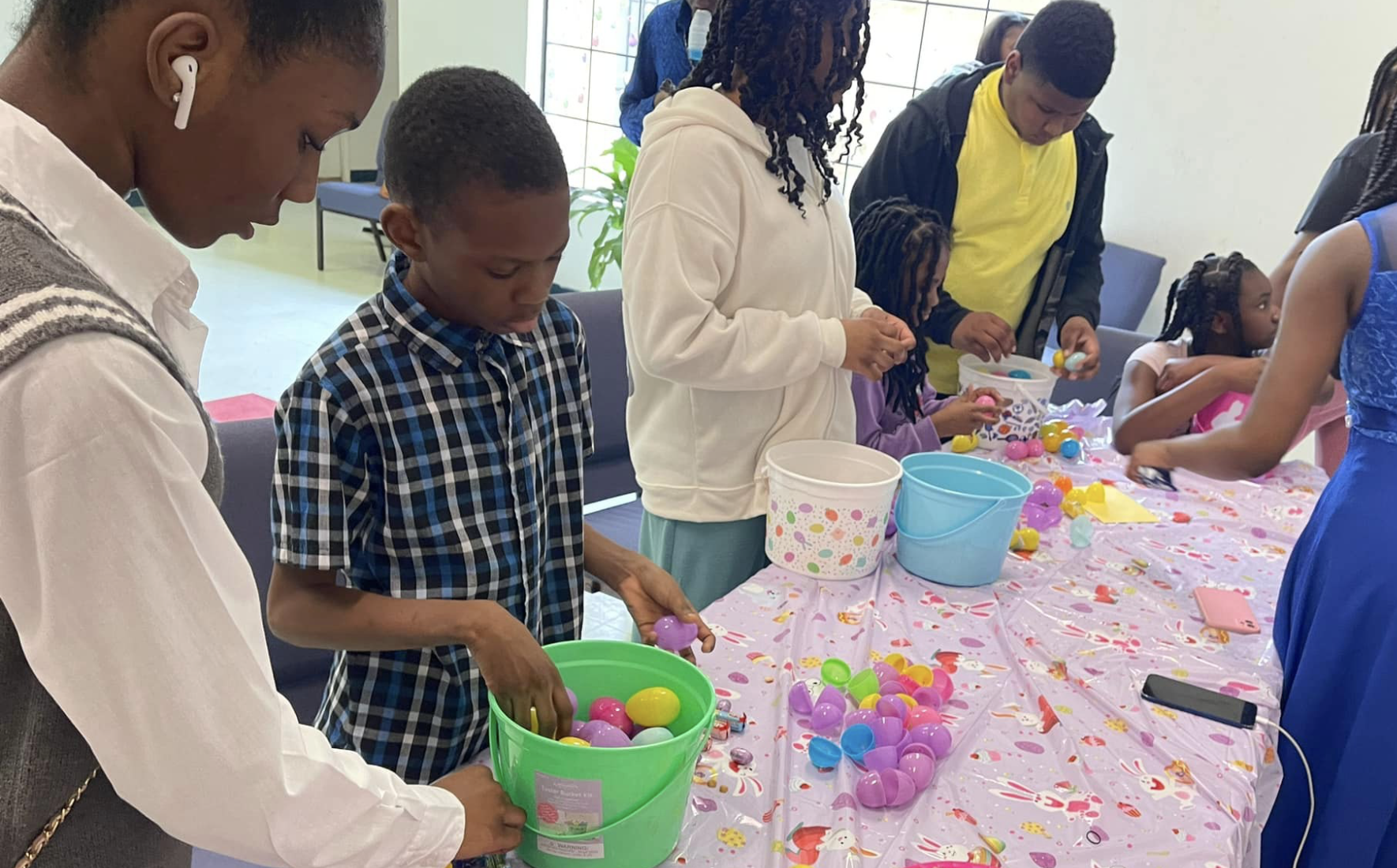 Children and adults decorating and filling plastic eggs with small toys and candies on a table covered with a pink Easter-themed tablecloth.