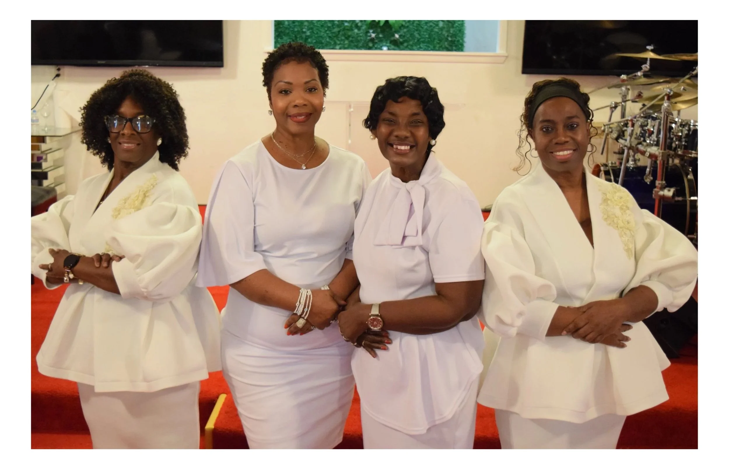 Four women dressed in white, standing together inside a church or event hall, with musical instruments visible in the background.