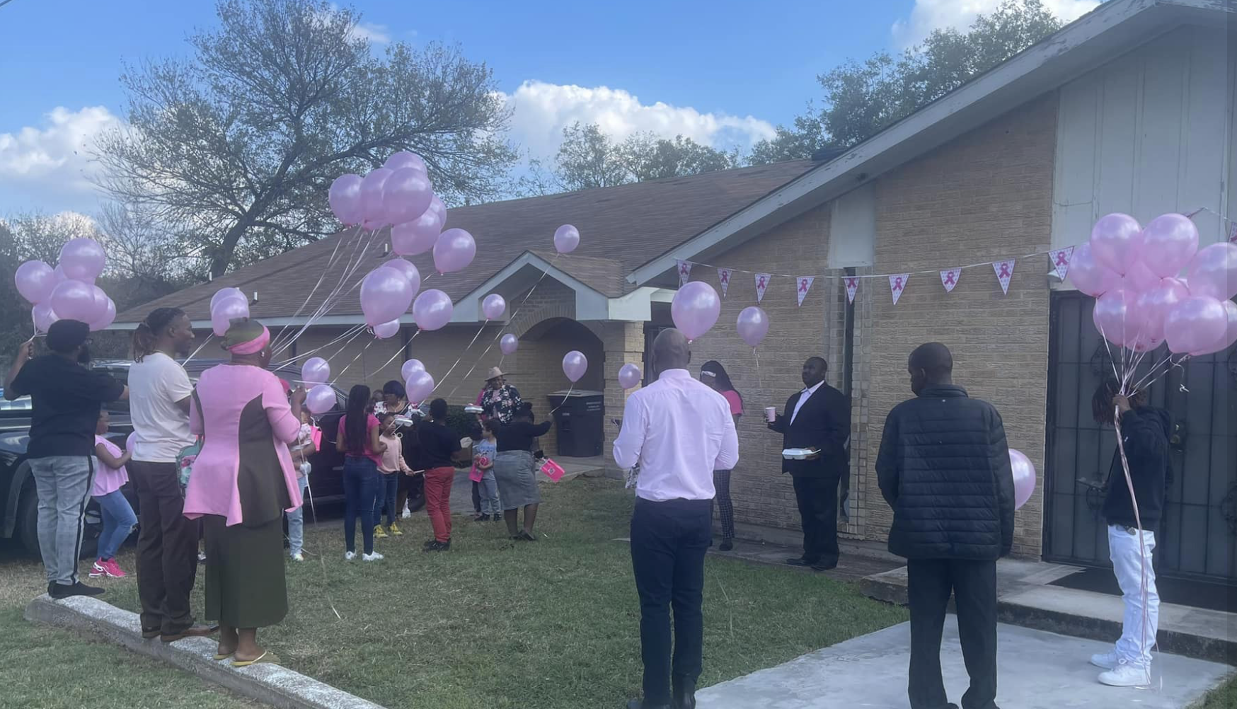 A gathering of people outside a house with purple balloons and pink ribbon banners for Breast Cancer Awareness.