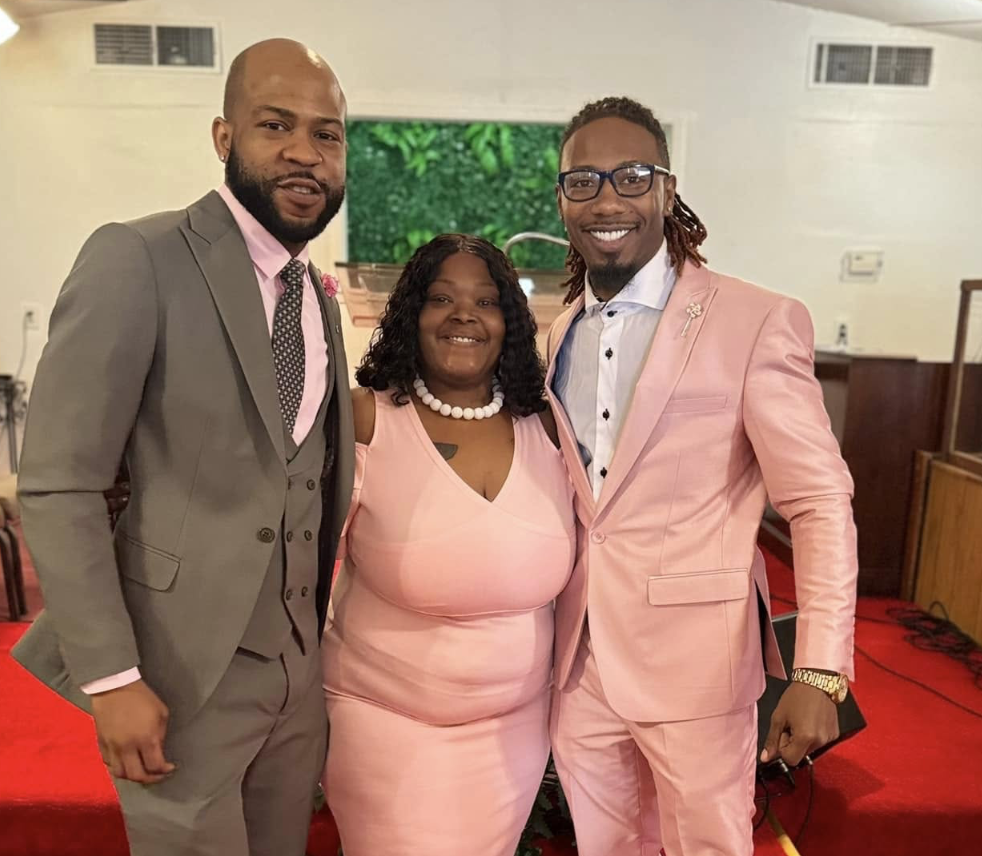 Three people dressed in formal attire posing together inside a church or hall, with a green leafy decoration background.
