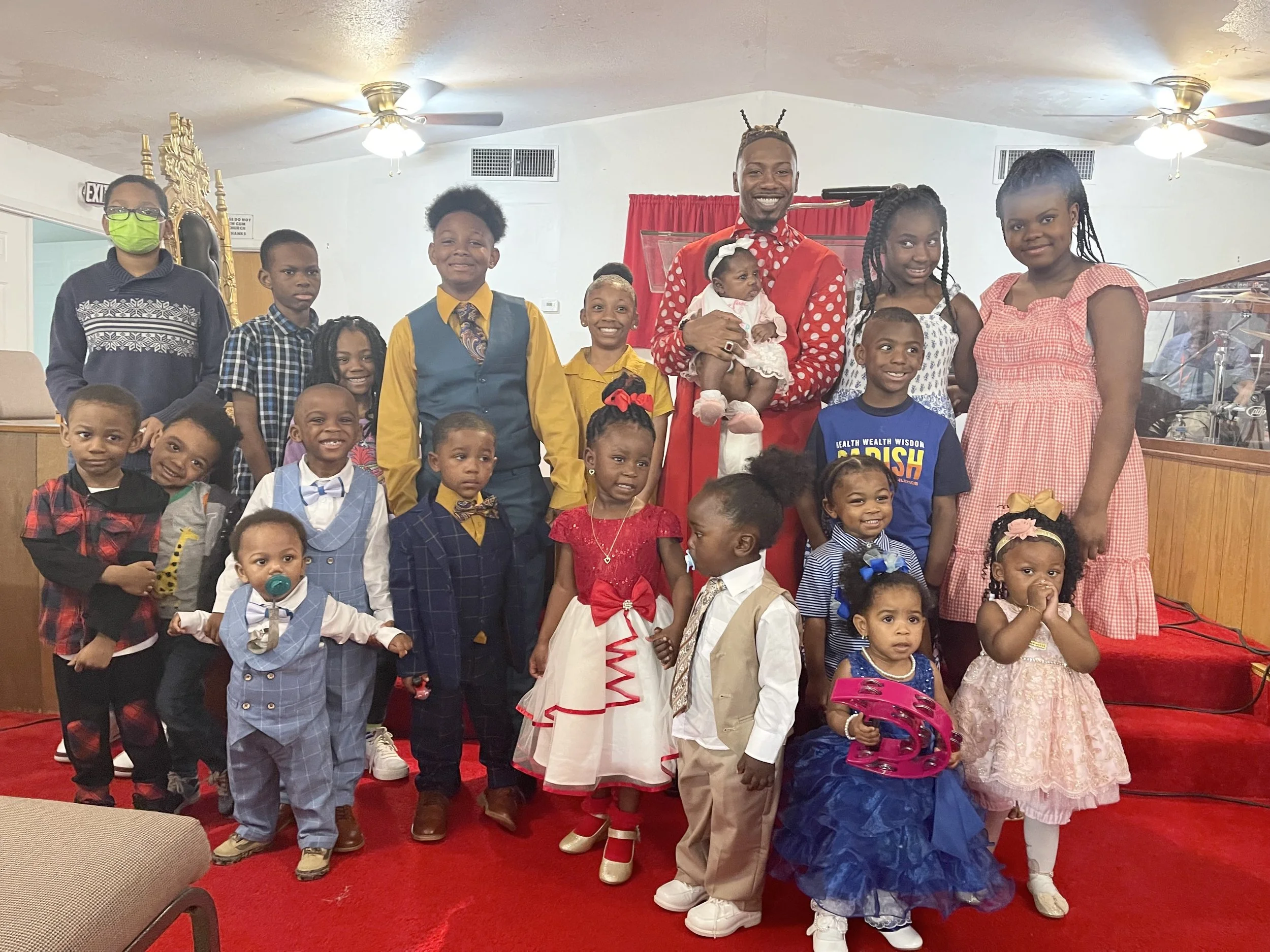 A group of children and adults, with many dressed in colorful outfits, posing together inside a church or community hall, around a man holding a baby. Some children are smiling and some are making different facial expressions.