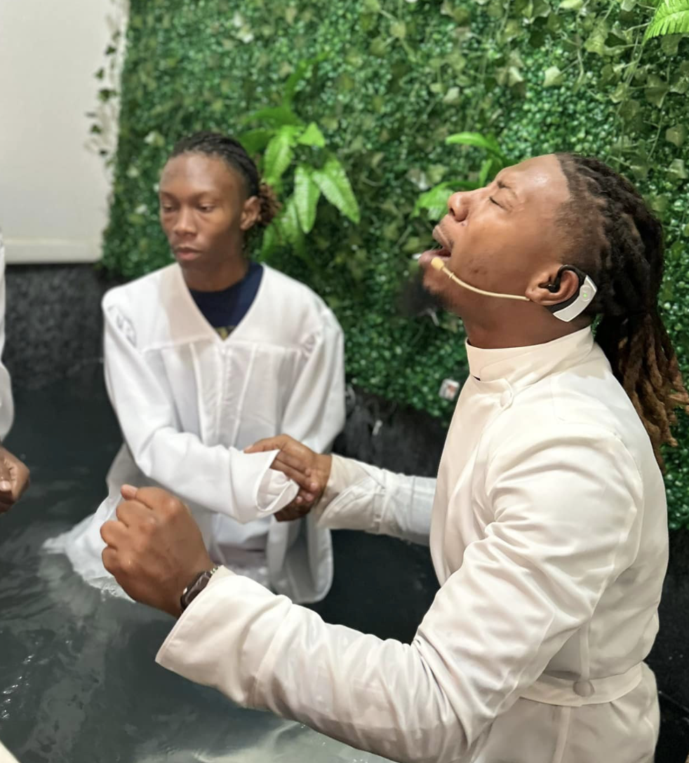 A man with dreadlocks and a headset, dressed in white, is being baptized in water by a young woman also in white. They are standing before a green leafy backdrop.
