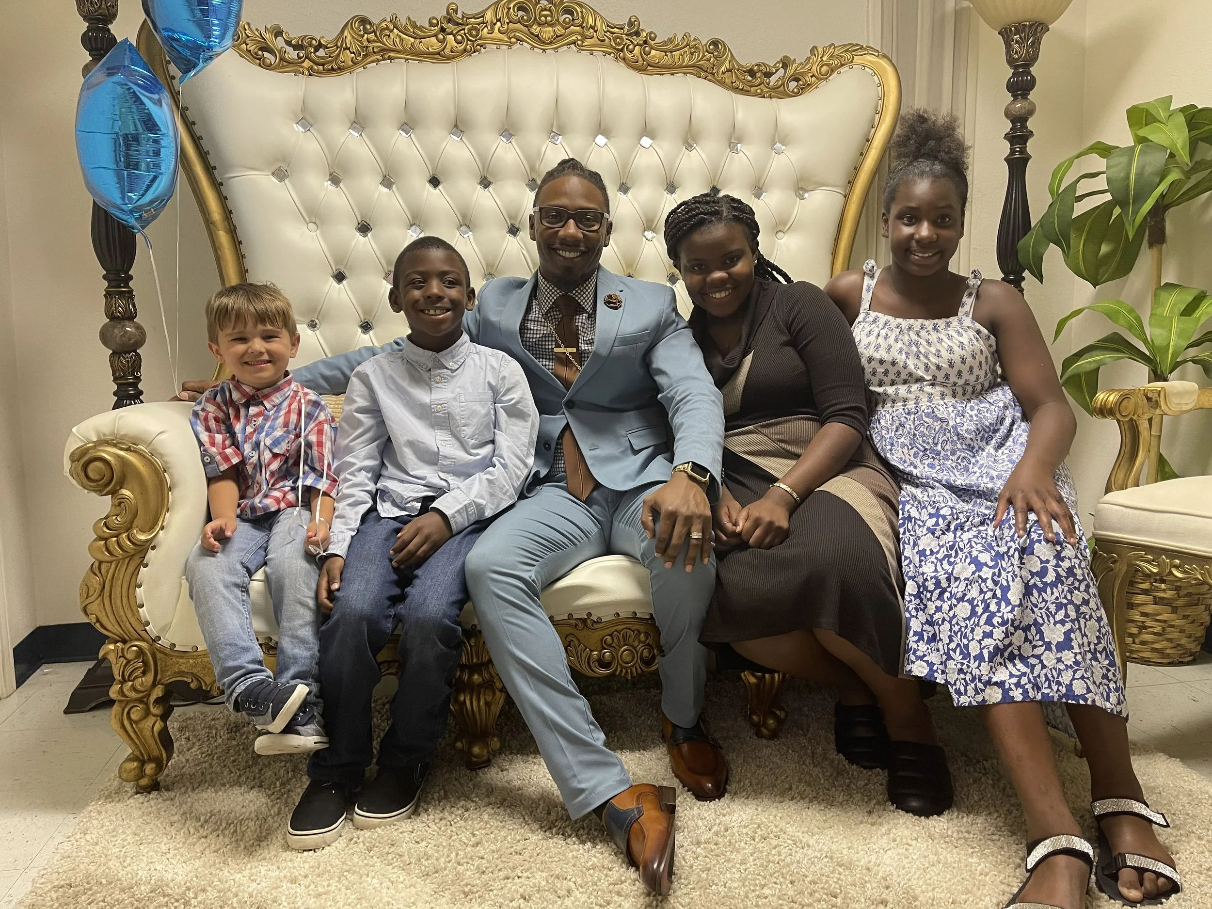 A group of five people, including a man in a light blue suit and four children, sitting on an ornate white and gold couch, with balloons and a potted plant in the background.