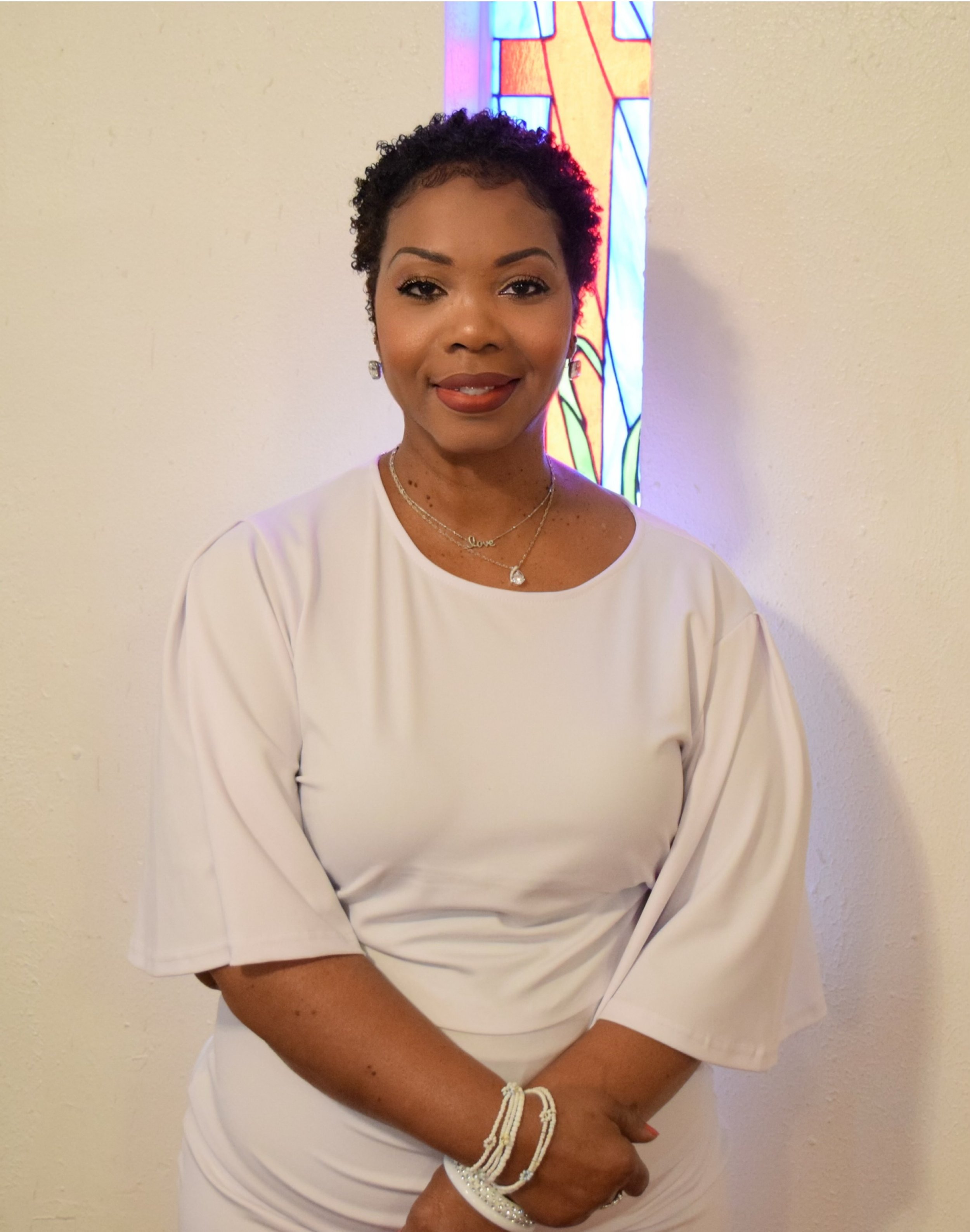 A woman with short curly hair, wearing a white dress and jewelry, standing in front of a beige wall and a stained glass window.