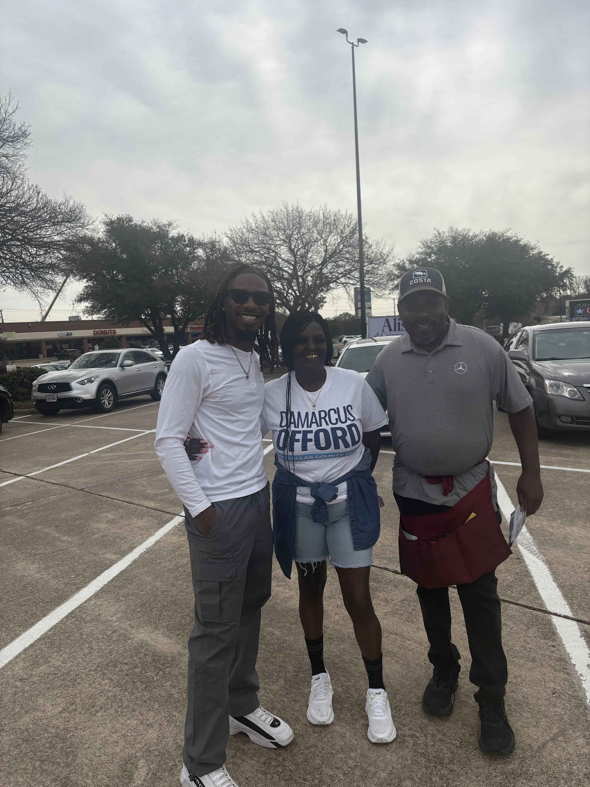 Three people standing in a parking lot, smiling at the camera on an overcast day. The woman in the middle wears a campaign T-shirt that reads "Damarcus G. Ford."