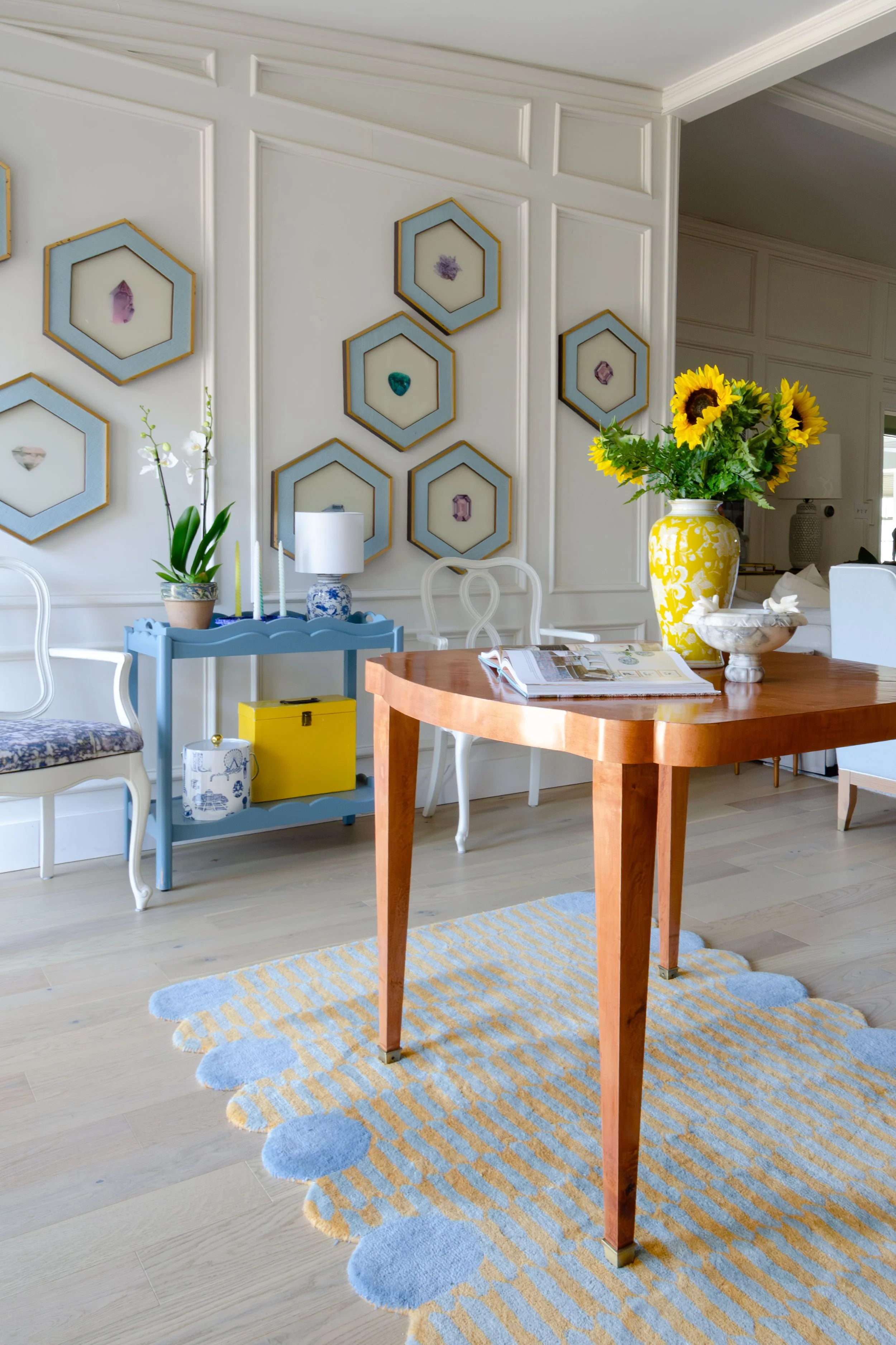 A dining room with a wooden table, a vase of sunflowers, a blue side table, white chairs, and a wall decorated with hexagonal framed decor.
