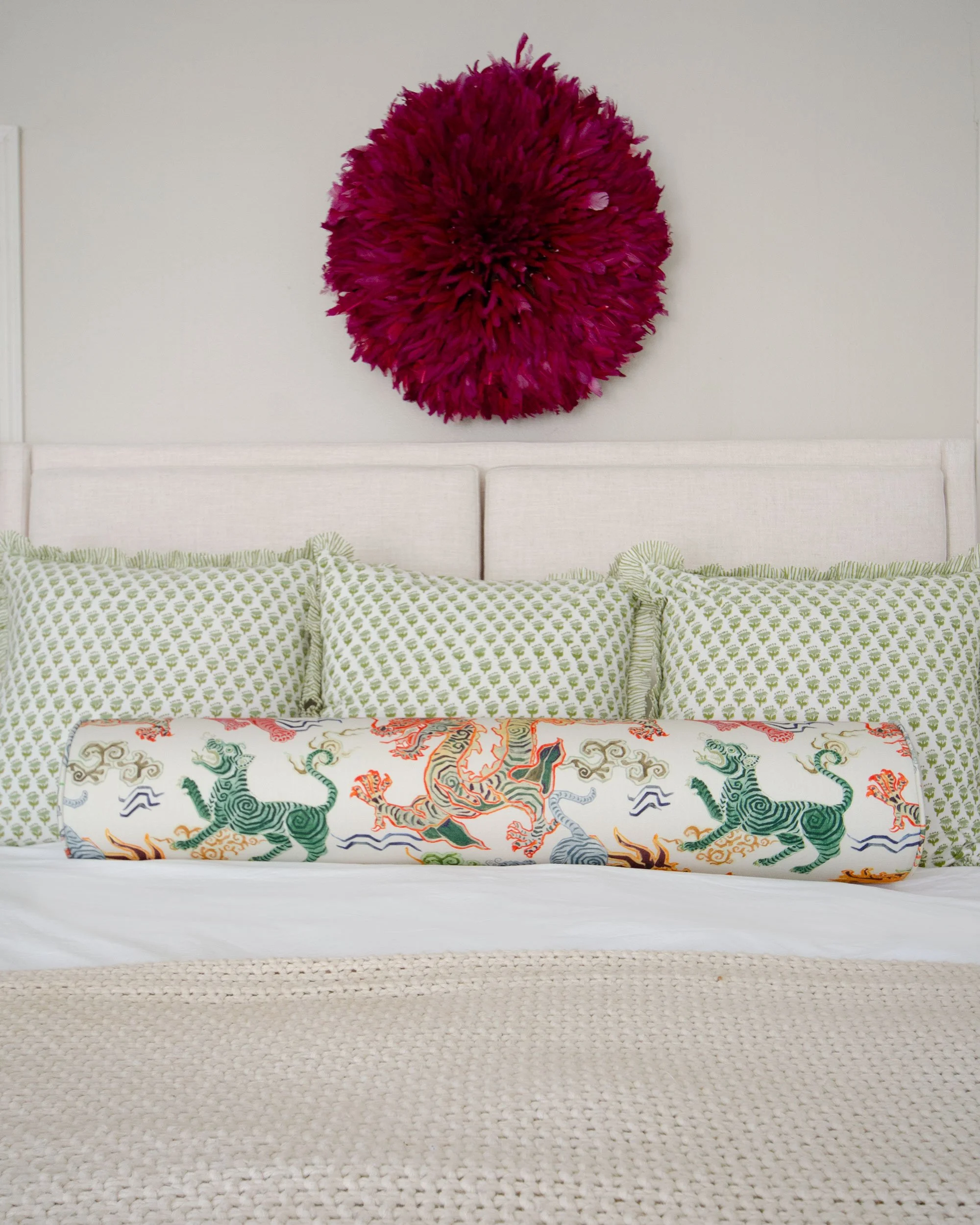 A bed with a white headboard, decorated with green and white patterned pillows, a colorful embroidered bolster pillow featuring zebras and lions, and a large, round, textured mauve wall art piece above.