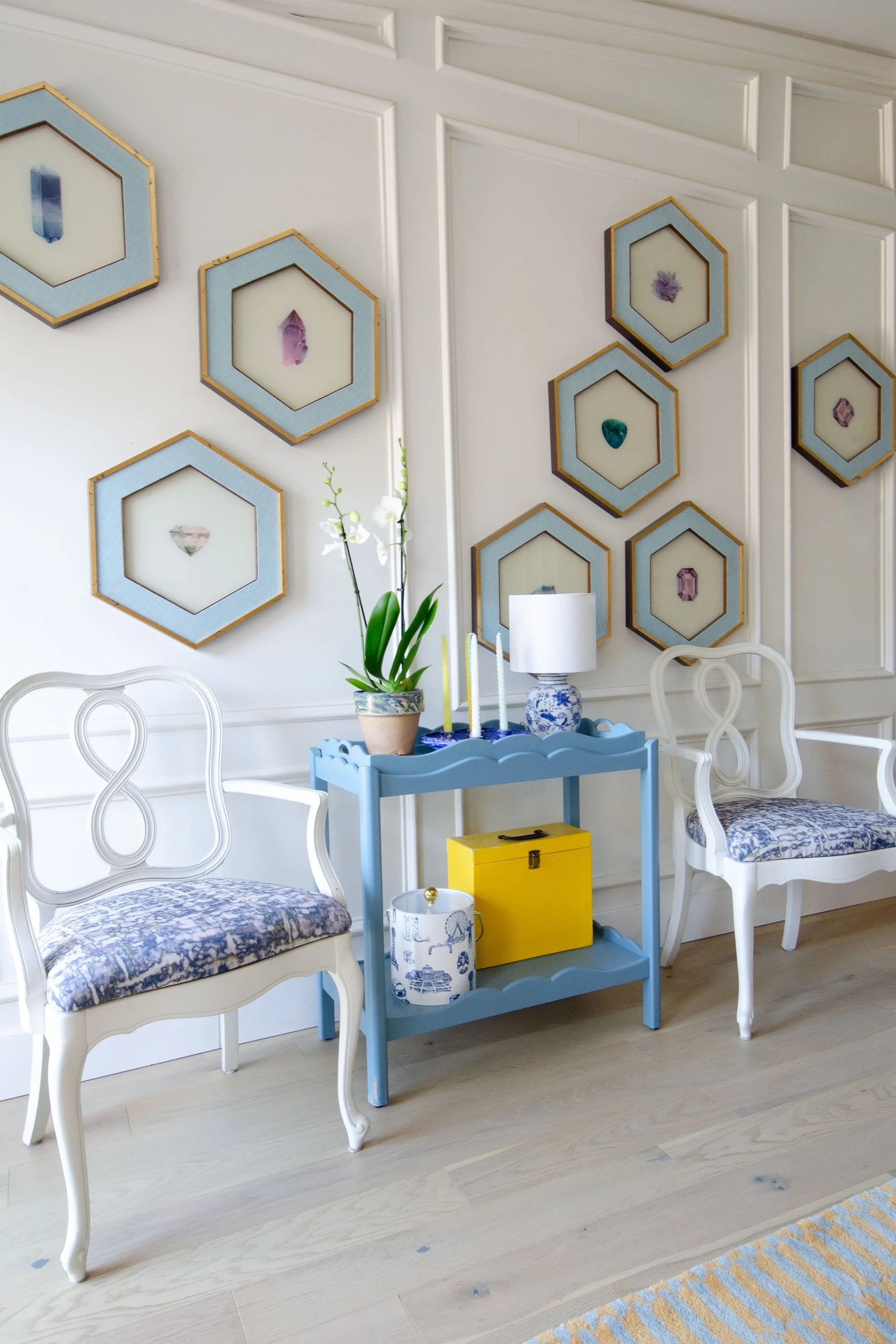 Decorative wall with framed art, a blue side table with a potted orchid, a table lamp, candles, a yellow box, and two white chairs with blue patterned cushions on a light wood floor.