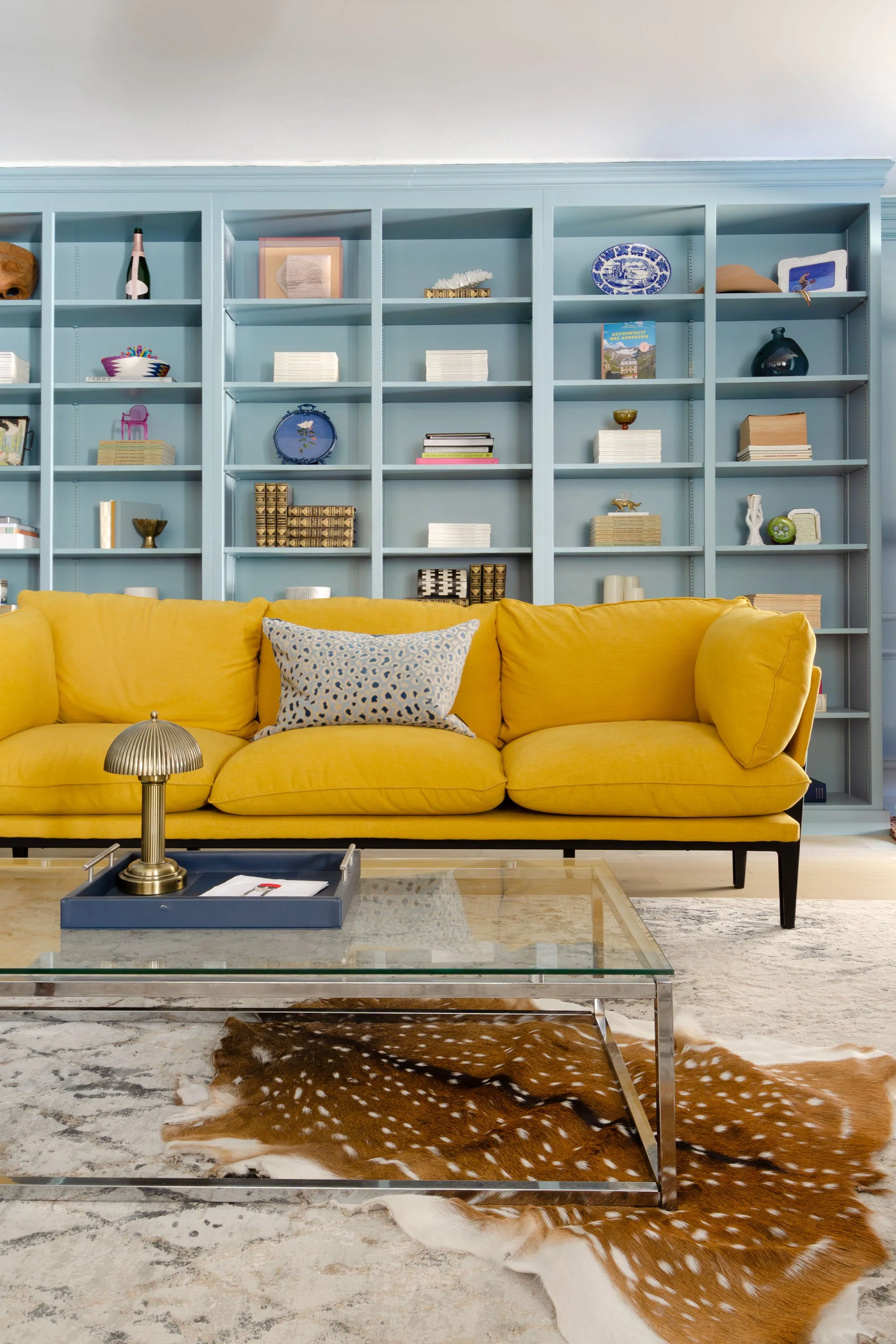 Living room with a yellow sofa, a glass coffee table with a brass lamp and a tray, a cowhide rug, and a blue built-in bookshelf filled with decorative items and books.