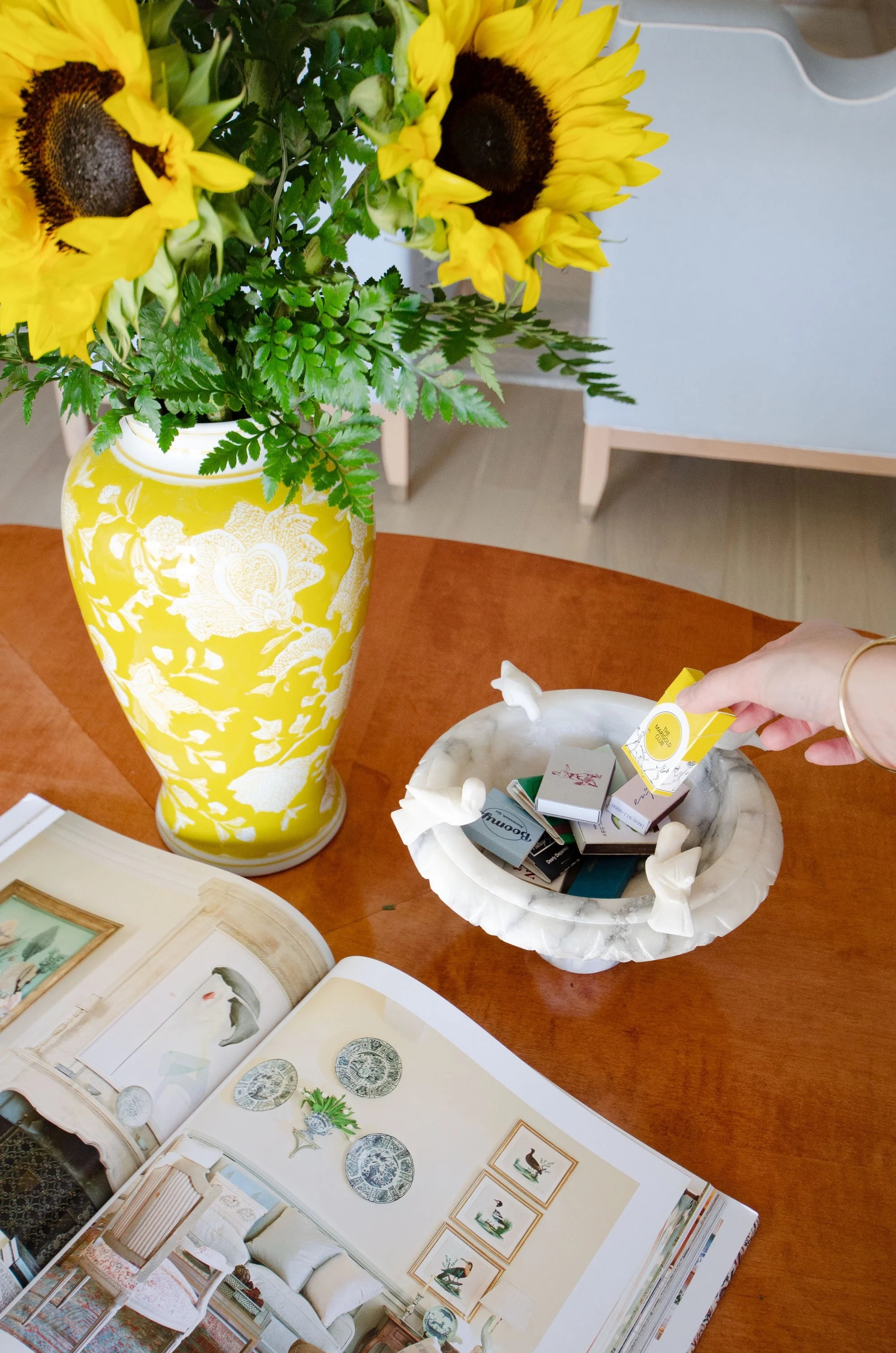 A yellow vase with white floral patterns holding a bouquet of yellow sunflowers and green fern leaves on a wooden table.