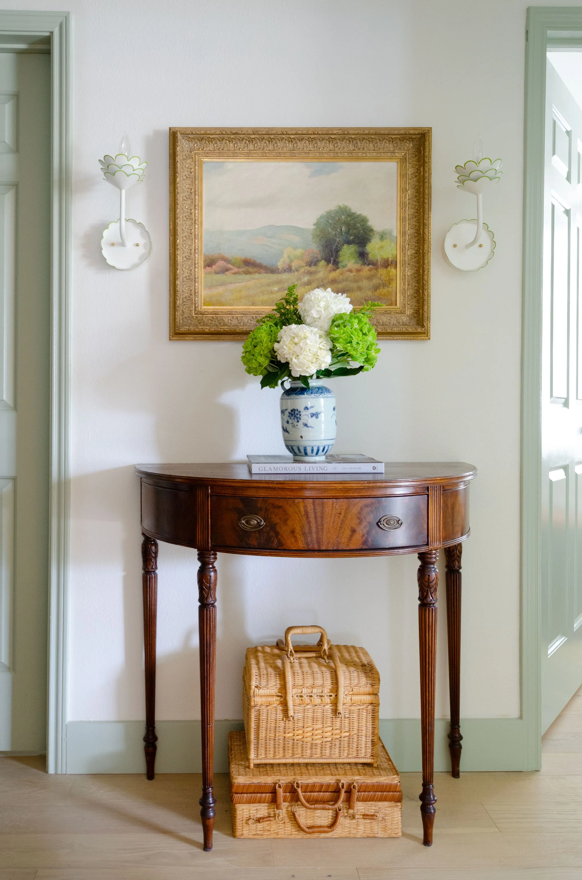 A wooden console table with a vase of green and white hydrangeas on top, decorative wall sconces, a framed landscape painting, and woven wicker baskets underneath.
