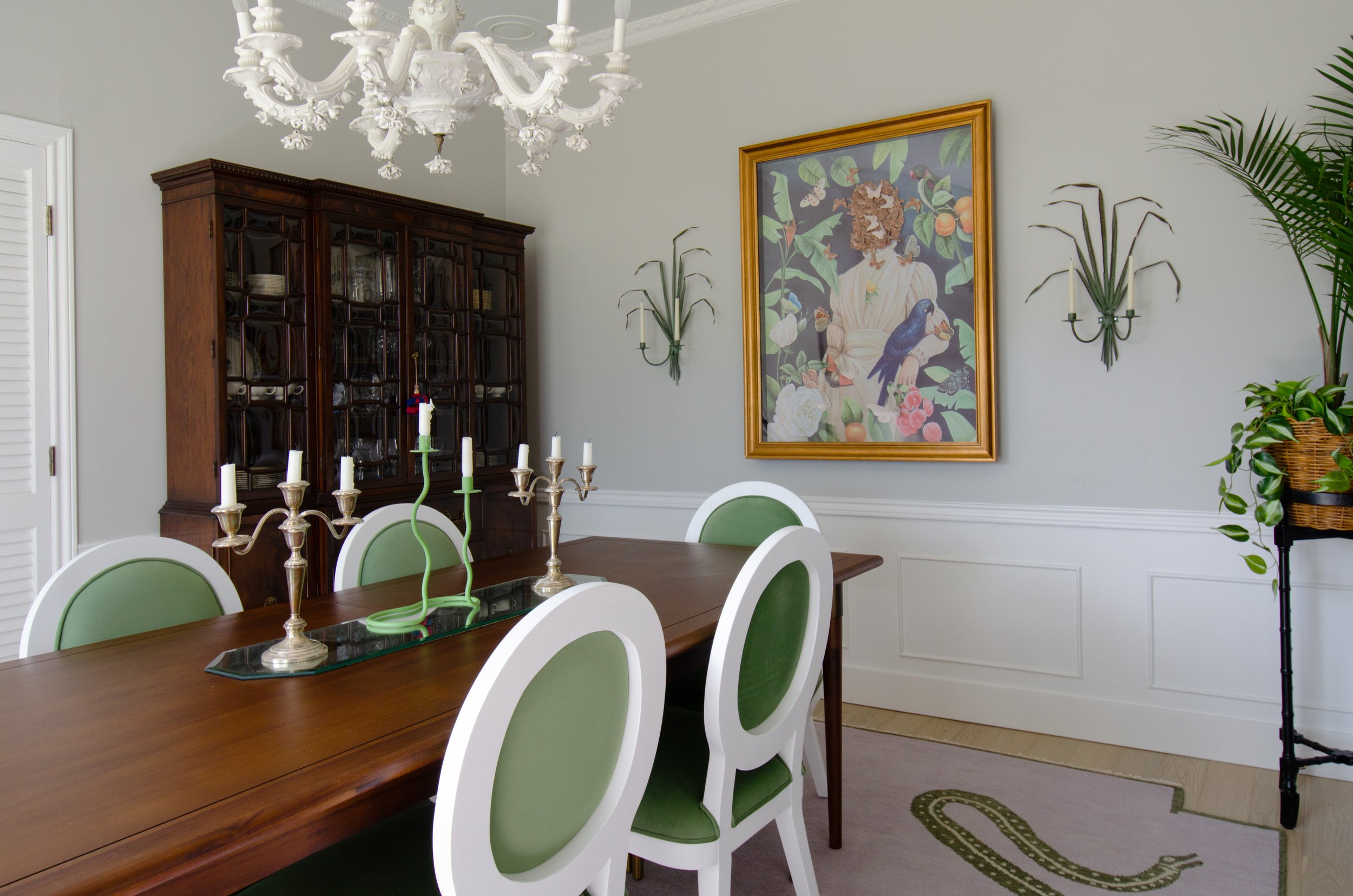 Dining room with a wooden table, green and white chairs, a glass and silver candelabra with candles, a dark wooden china cabinet, a painting of a person with a lion's face and surrounded by leaves, green plants in the corners, and a white chandelier 