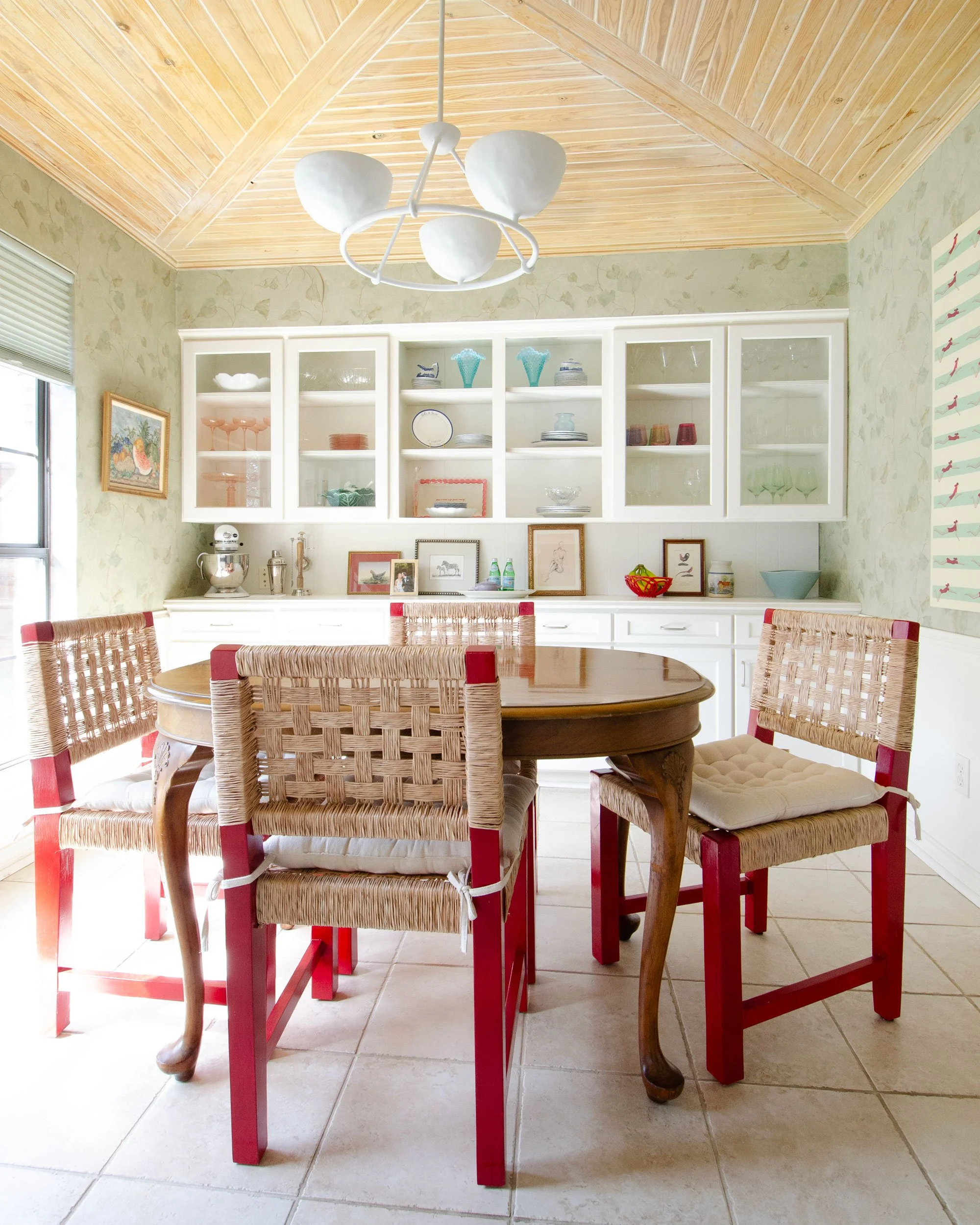 A dining room with a round wooden table, four wicker chairs with red-painted wooden legs, and built-in white cabinets filled with glassware and decorative items. The room has a wood-paneled ceiling, a chandelier, and a window with blinds.