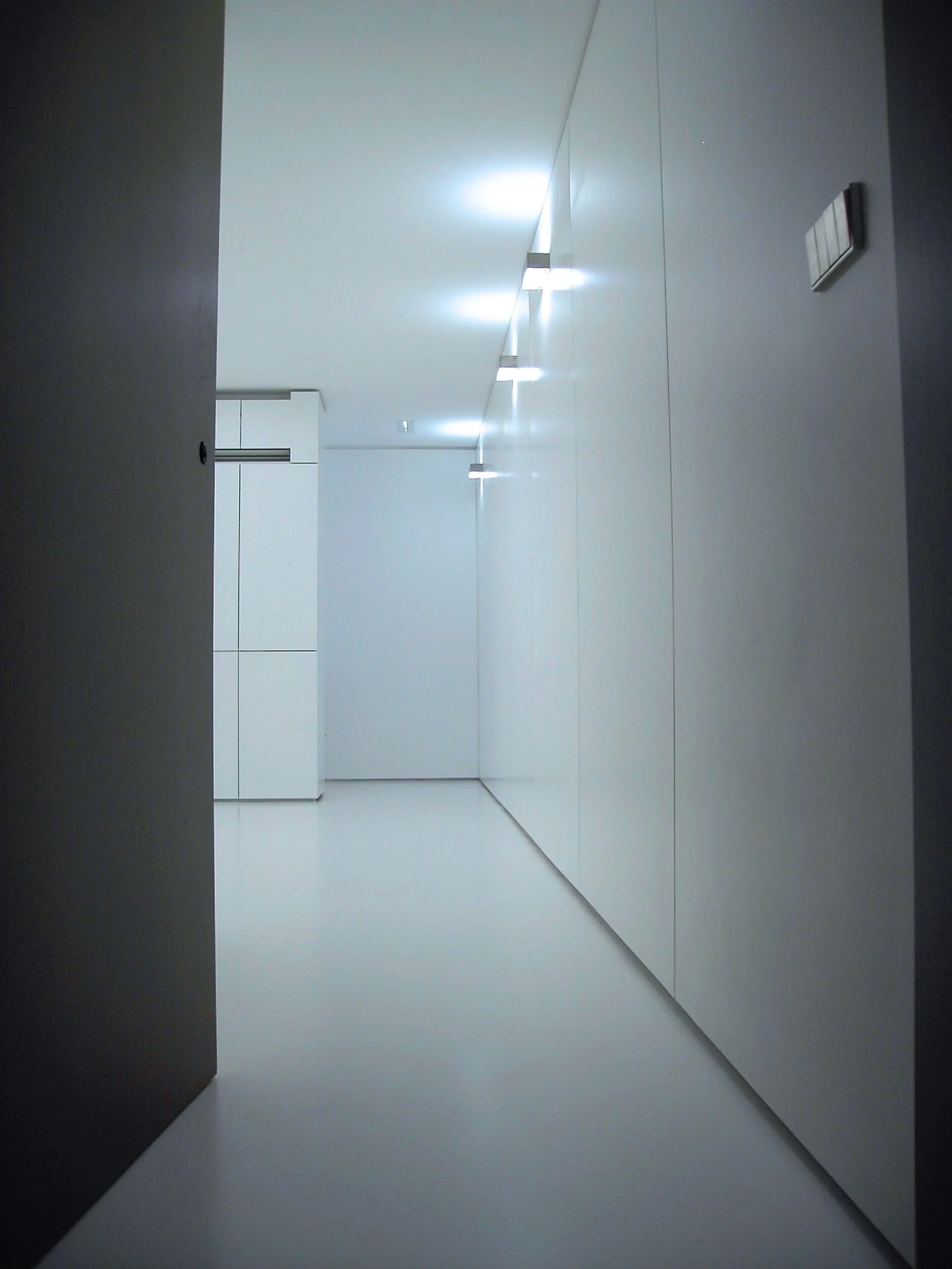 Corredor de interior com paredes e teto brancos, piso branco, iluminação de luz branca, armários brancos na parede ao fundo.