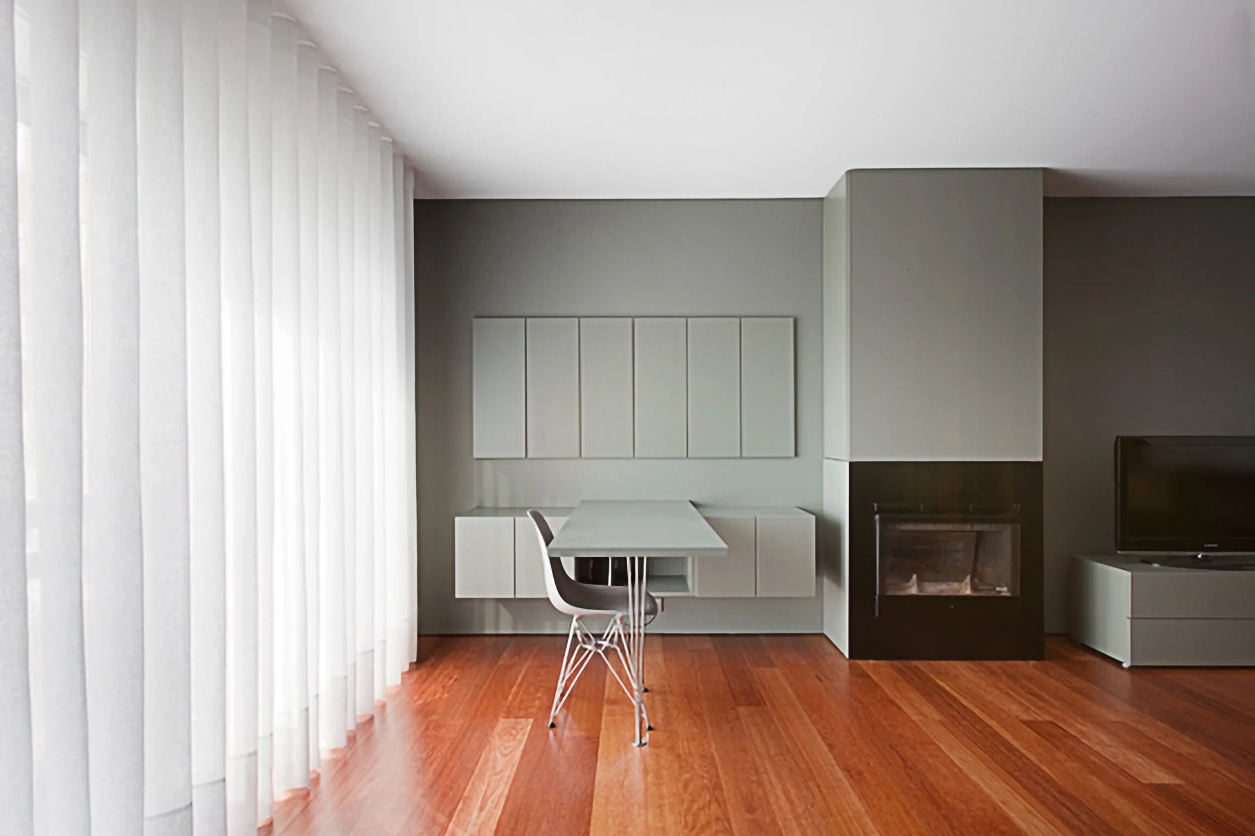 Sala moderna com parede cinza, janela com cortina branca, mesa de jantar e cadeira, lareira preta, televisão e móvel cinza, piso de madeira.