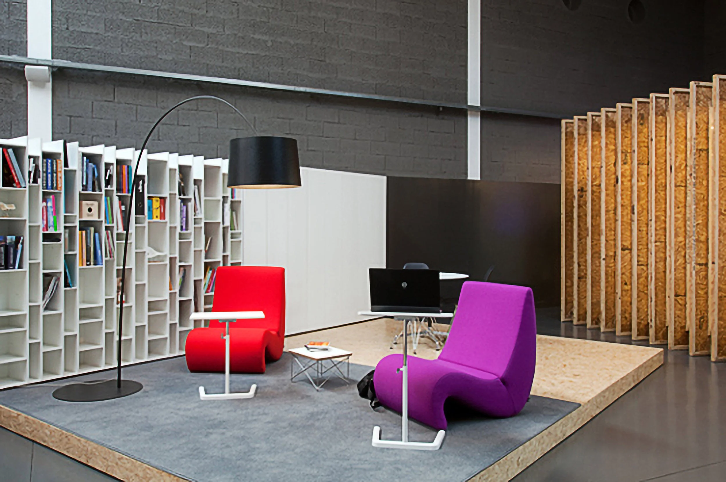 Espaço de coworking com cadeiras coloridas, biblioteca de estantes brancas, mesa de escritório com computador, e paredes de concreto.