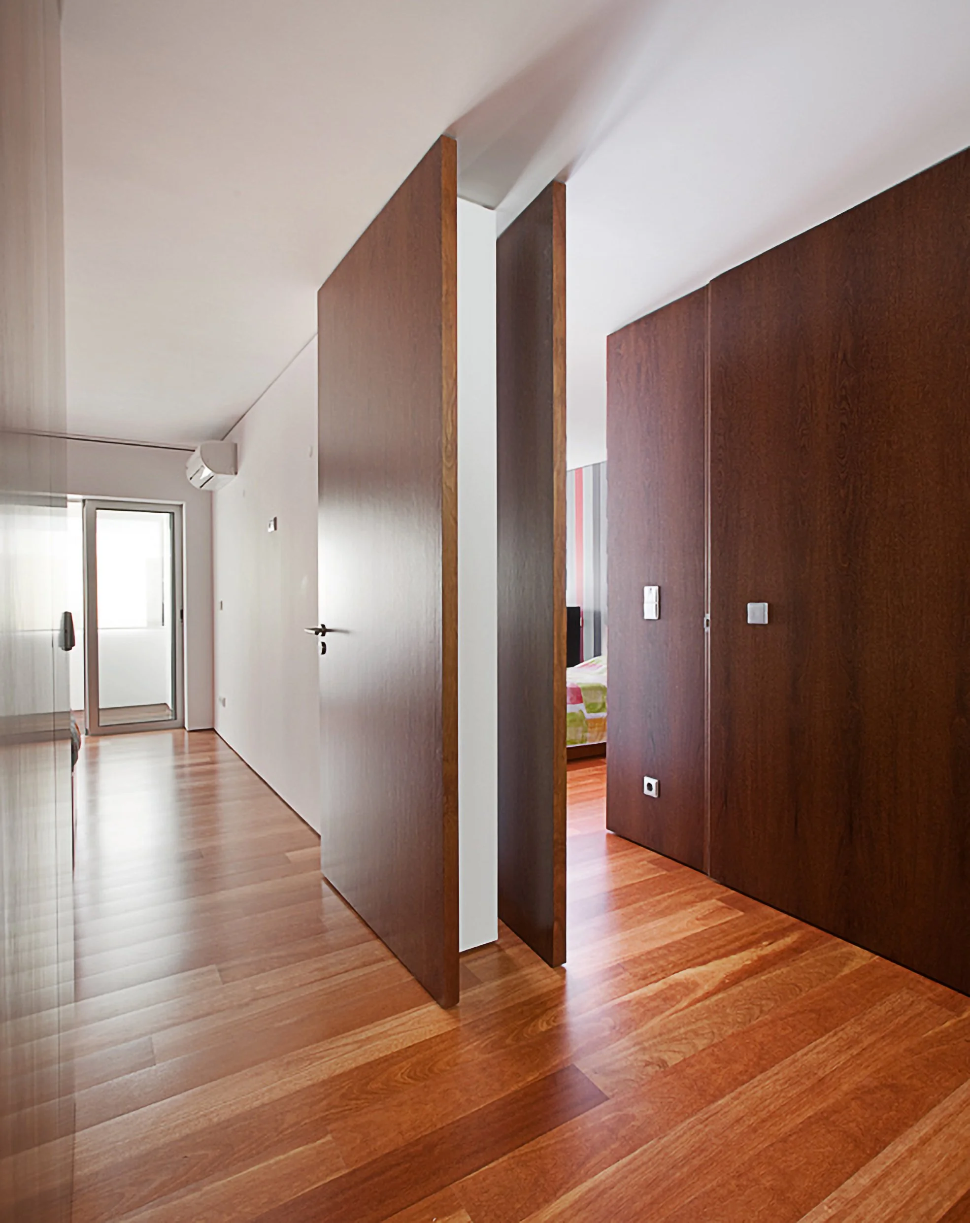 Corredor com portas de madeira que levam a quartos, piso de madeira, parede branca e janela na extremidade.