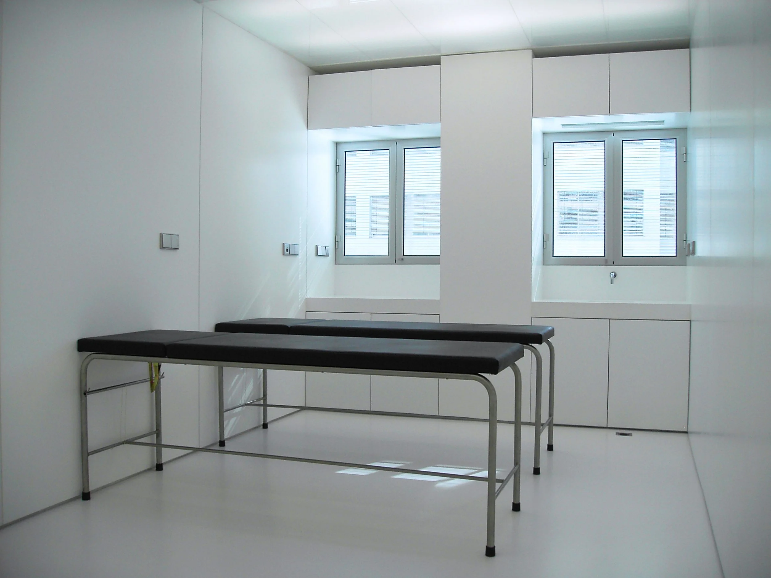 Sala de exames ou consultório com duas macas pretas, paredes brancas, janelas grandes com persianas, armários brancos e iluminação natural.