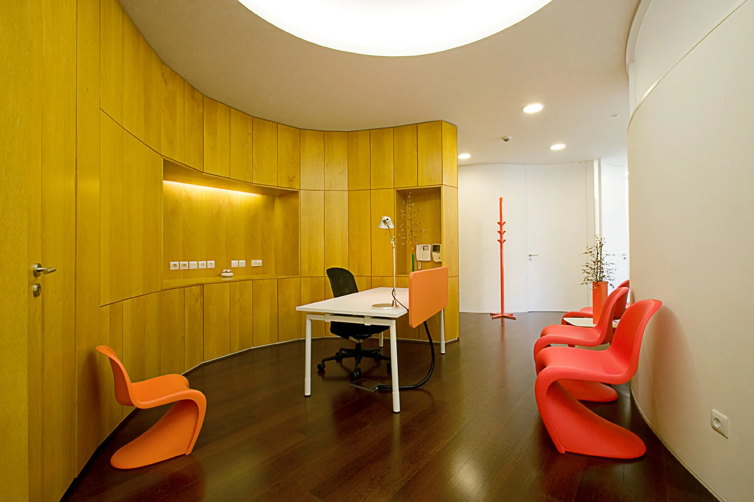 Sala de reunião com paredes de madeira, mesa branca, cadeira preta na frente e cadeiras rosa ao lado, com iluminação suave