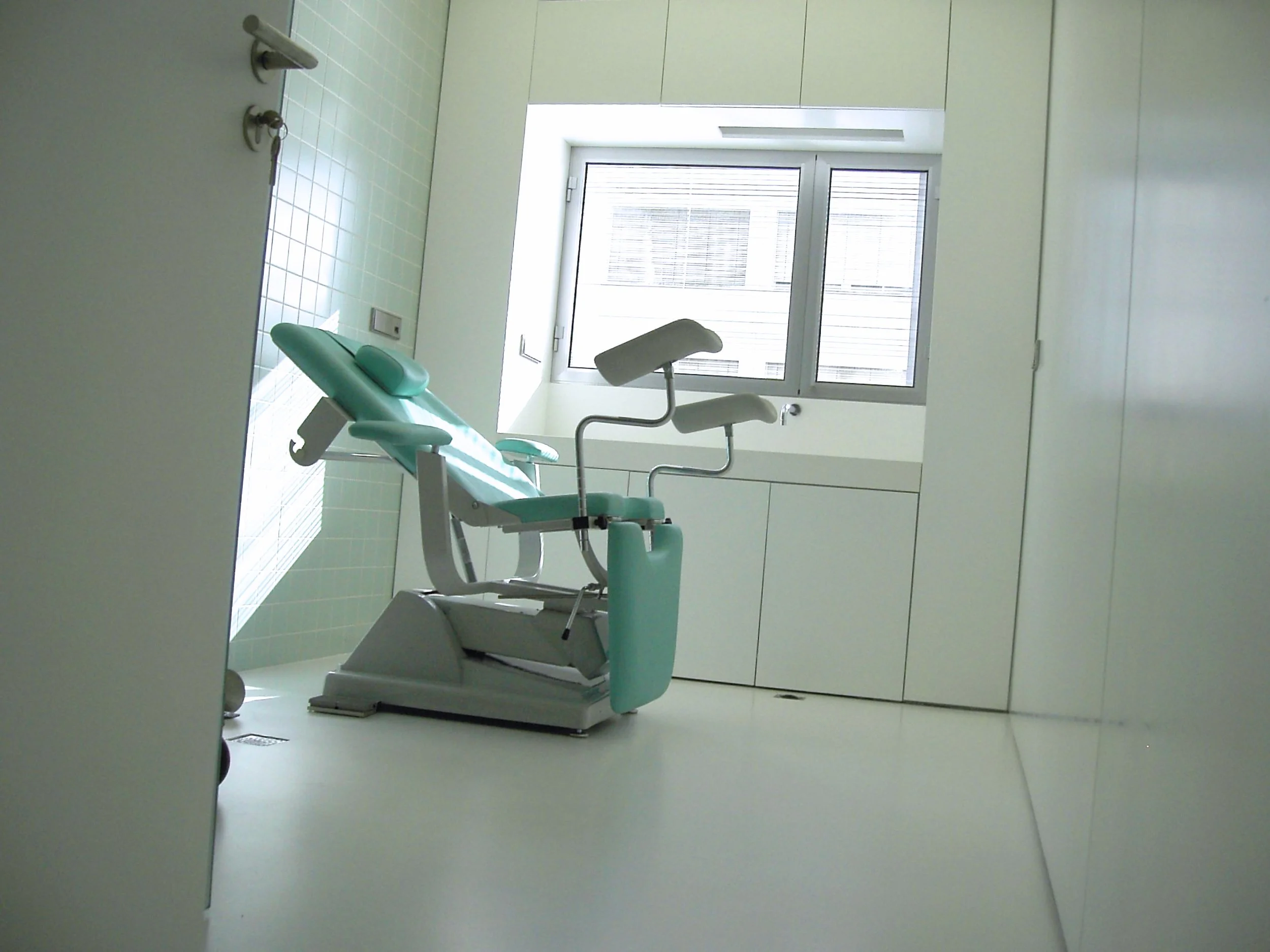 Configuração de uma cadeira odontológica em uma sala clínica bem iluminada, com janela grande ao fundo, paredes brancas e armários embutidos.