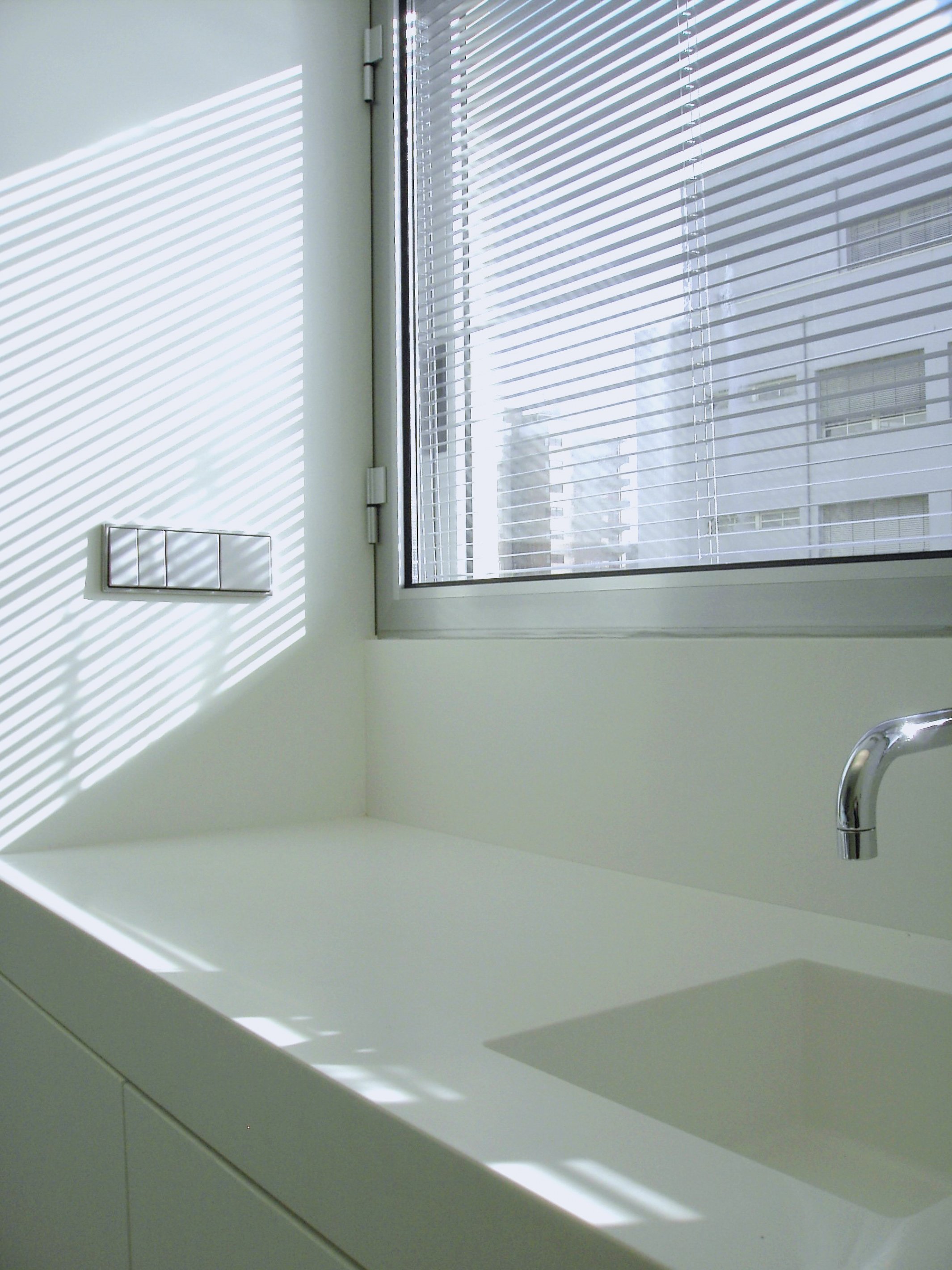 Janela com persianas de metal deixando a luz do sol entrar, refletindo sombras na pia branca.