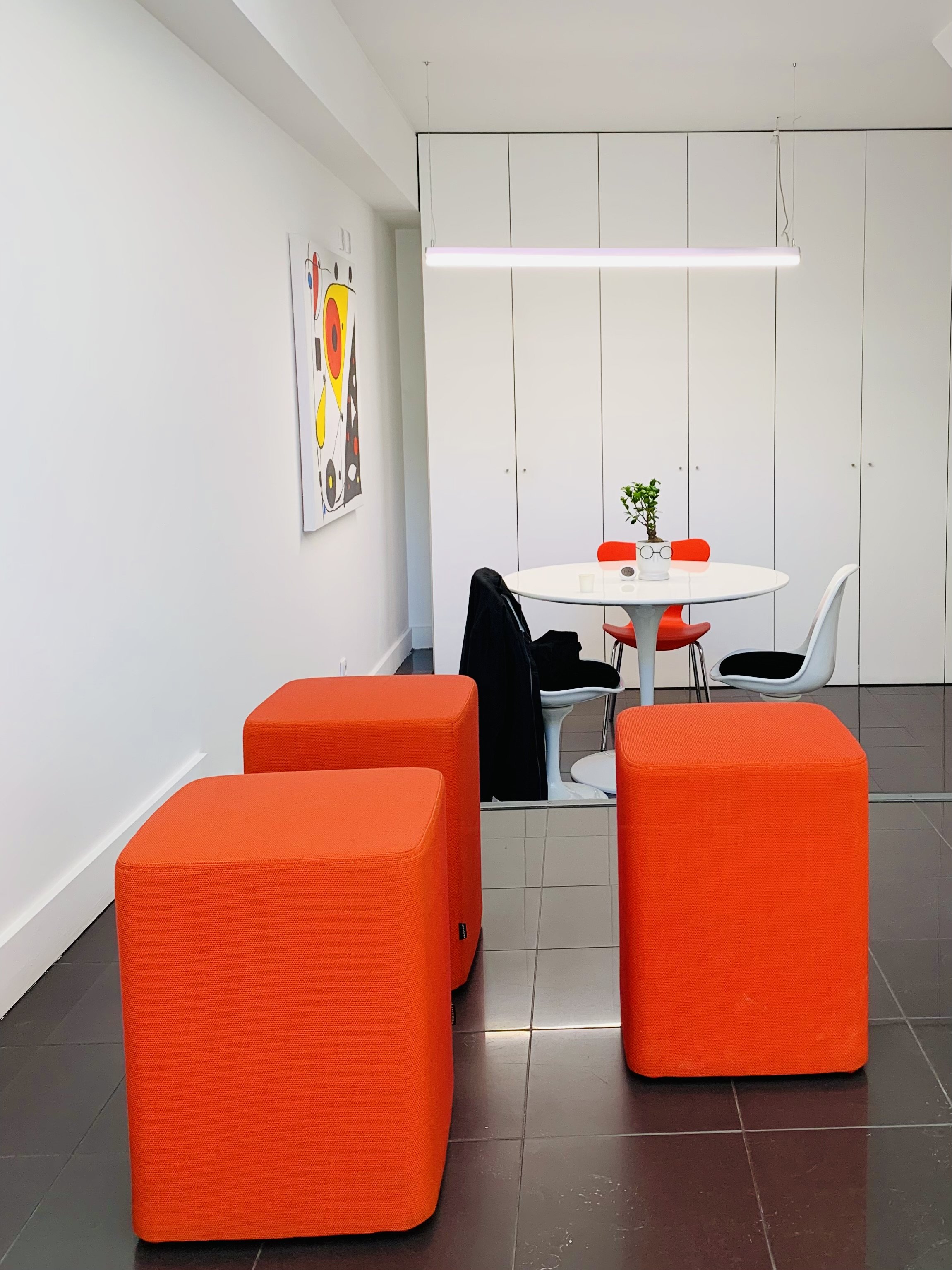 Sala de reunião moderna com duas poltronas brancas ao redor de uma mesa branca redonda, uma planta em vaso na mesa, quadro colorido na parede branca, cabine de vidro com revestimento espelhado e estantes brancas no fundo. Na frente, três pufes laranja.