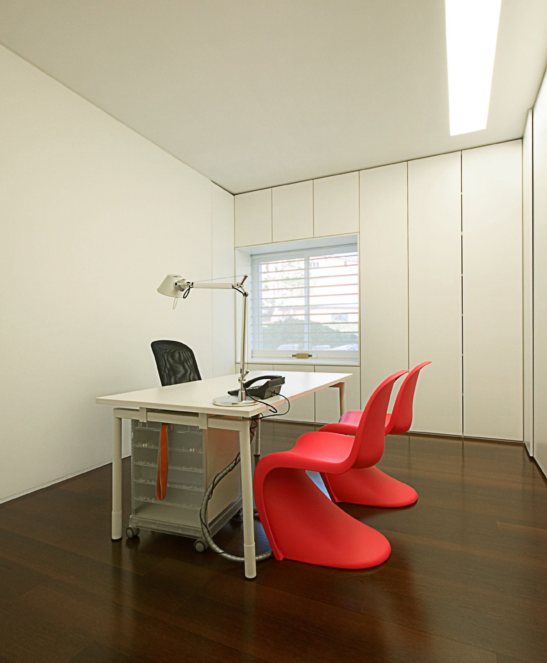 Escritório minimalista com mesa branca, cadeira de escritório preta, duas cadeiras vermelhas com design moderno, janela com persianas, piso de madeira escura e paredes brancas.