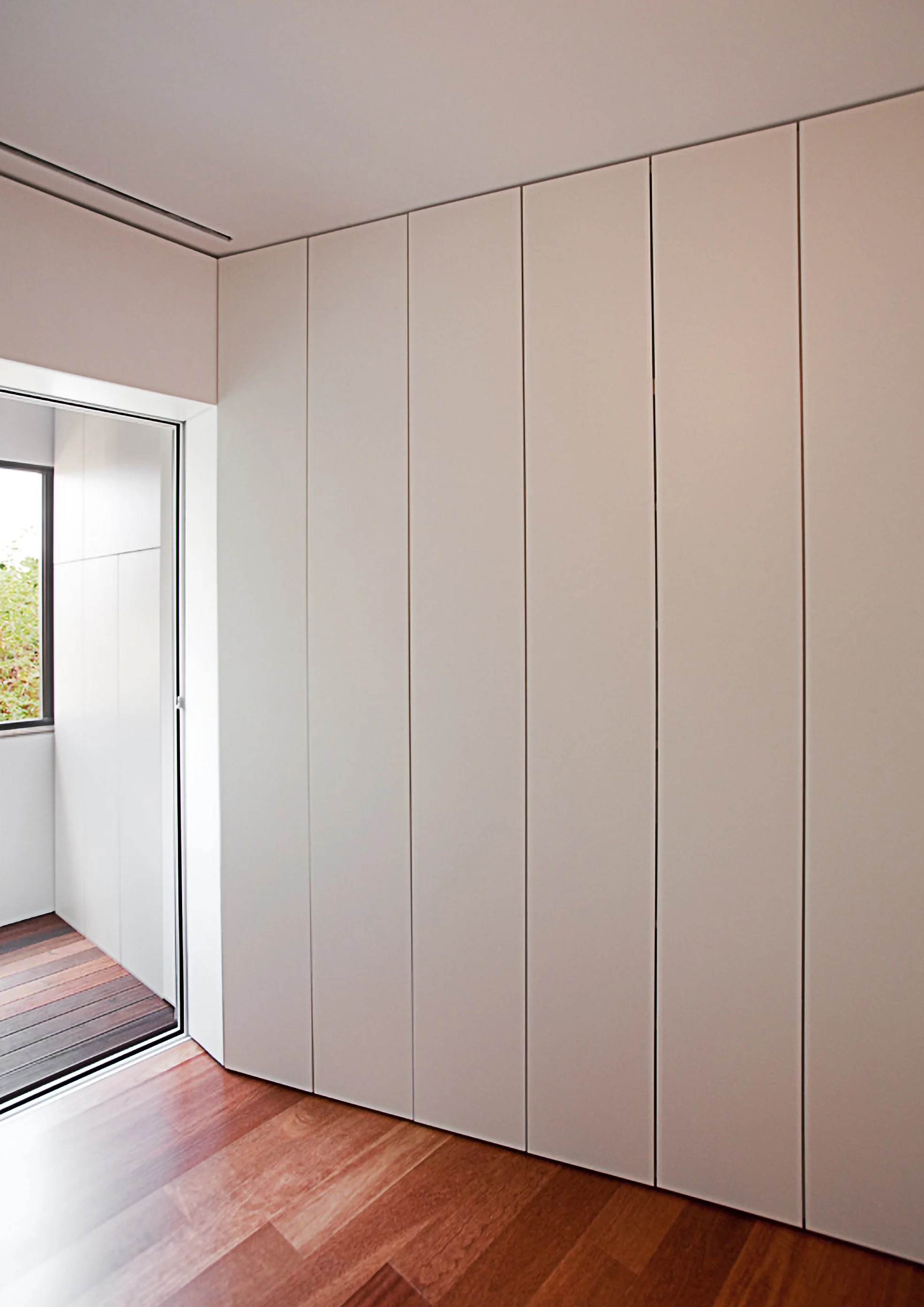 Parede branca com portas de armário e piso de madeira, com janela ao lado.
