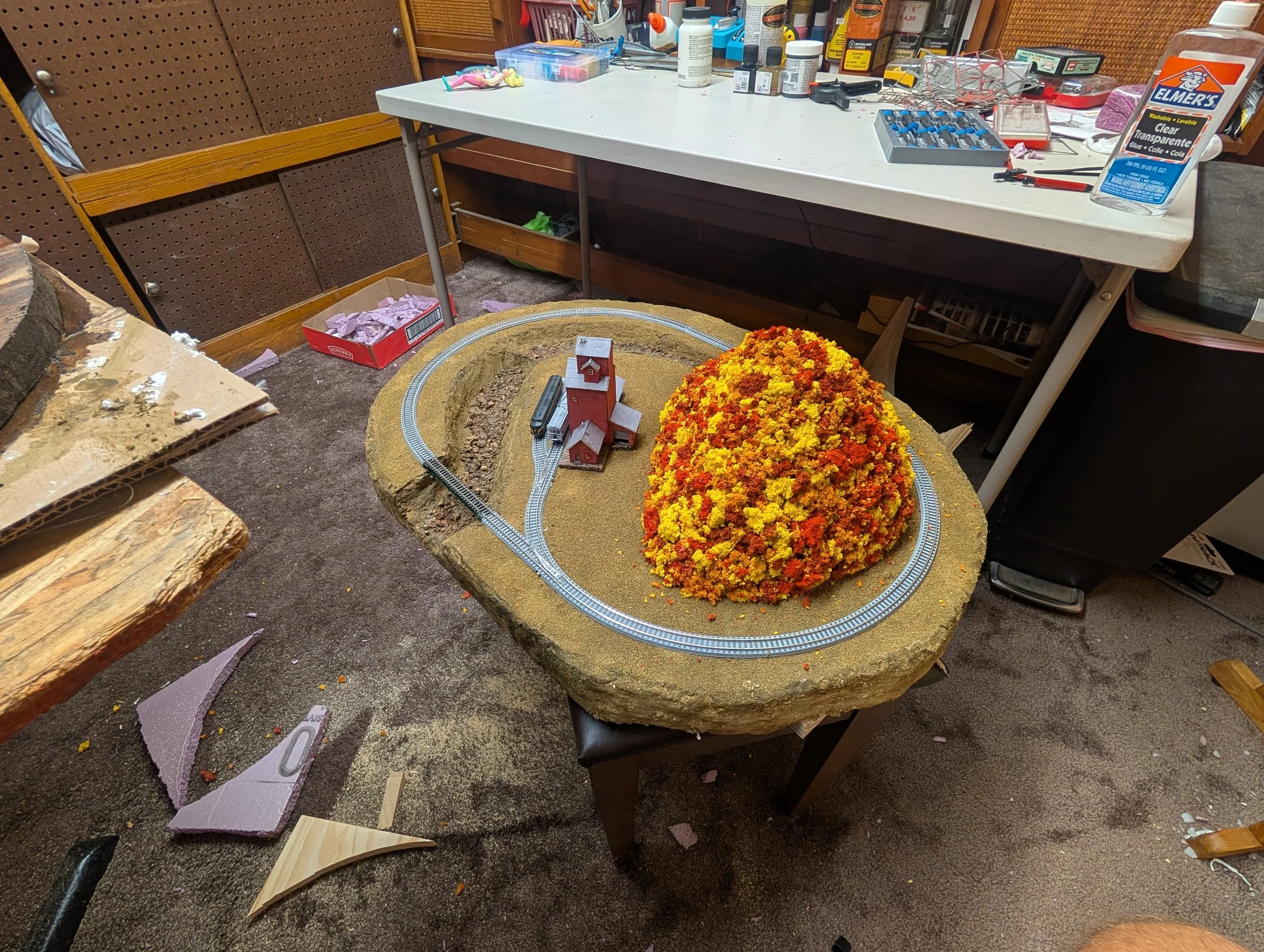 A model train set on a circular track with a miniature house and a large orange, yellow, and red artificial bush or tree on a miniature landscape.