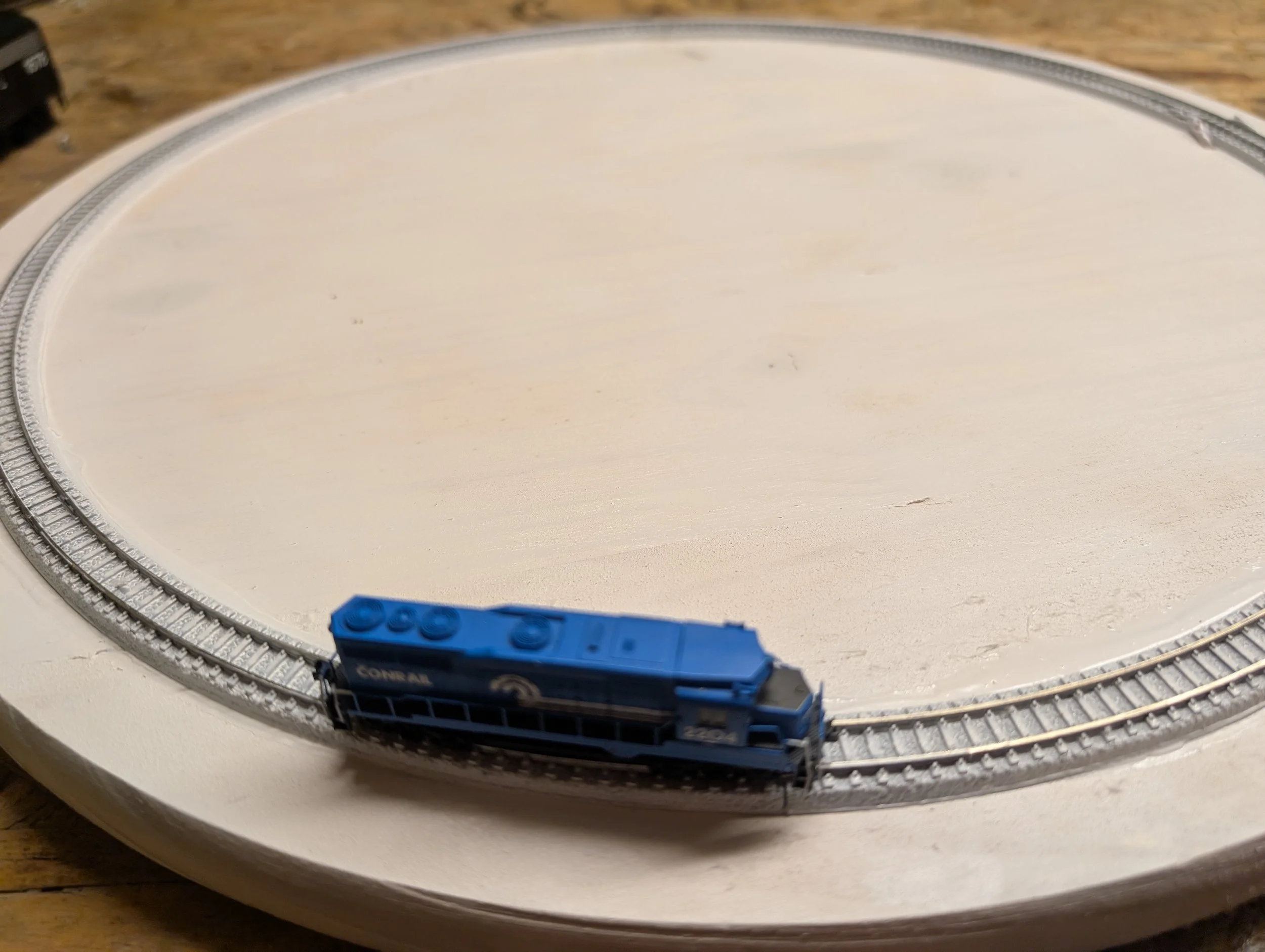 Small blue toy train on a circular train track, with a plain white surface and a wooden background.