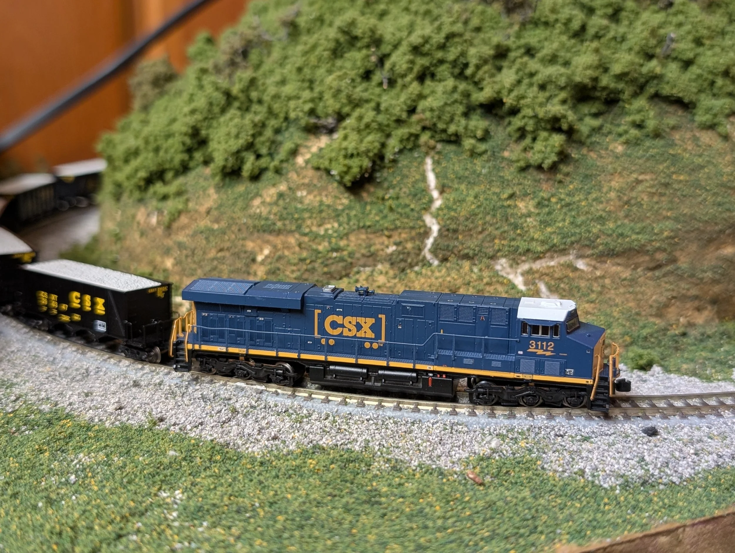 A detailed model train locomotive on a miniature track. It is painted blue with yellow accents, labeled with 'CSX' and the number '3112'. The background features a landscape with green hills and a small creek.