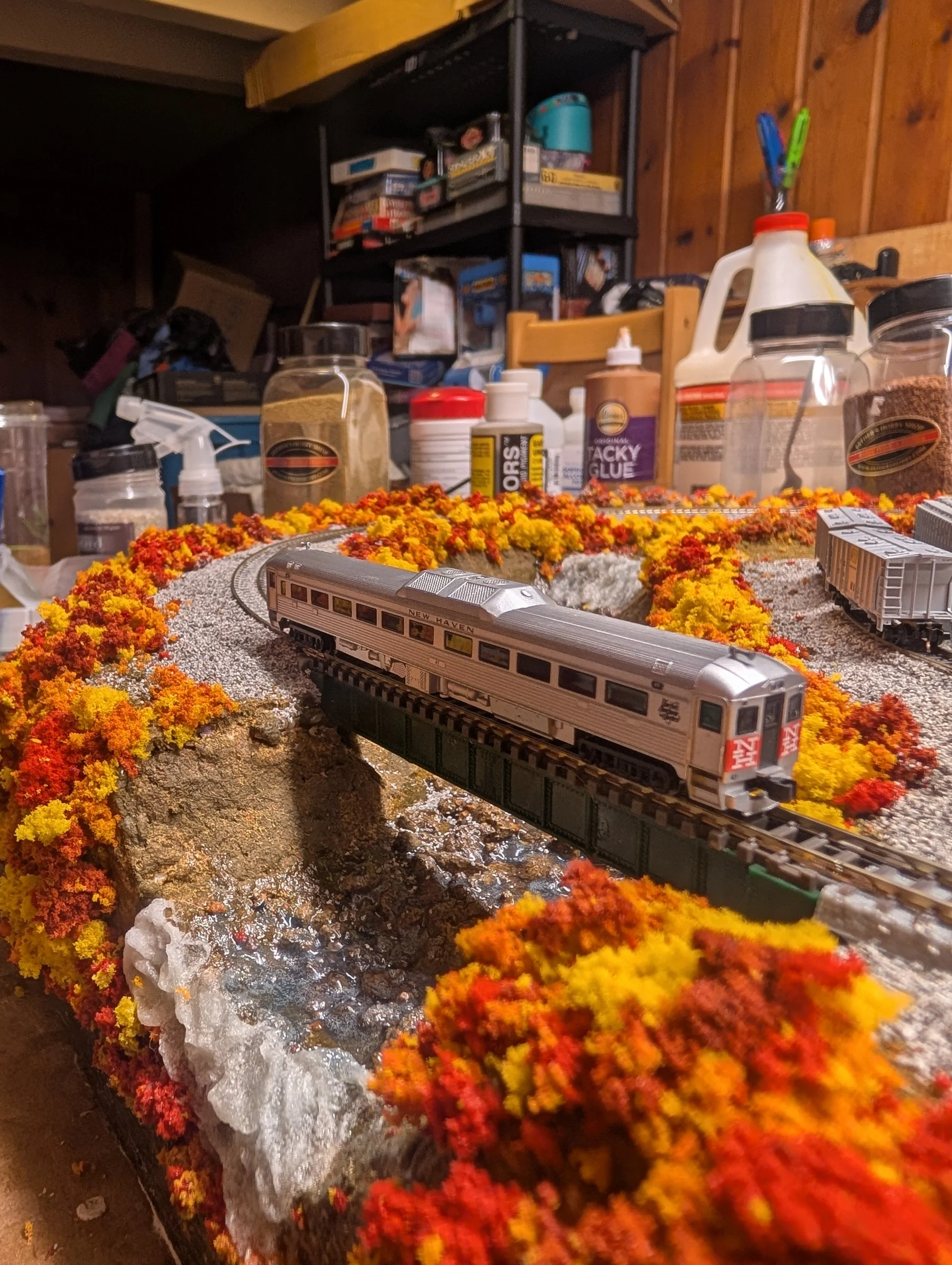 Model train set inside a room with shelves and various supplies, decorated with colorful moss and foliage.