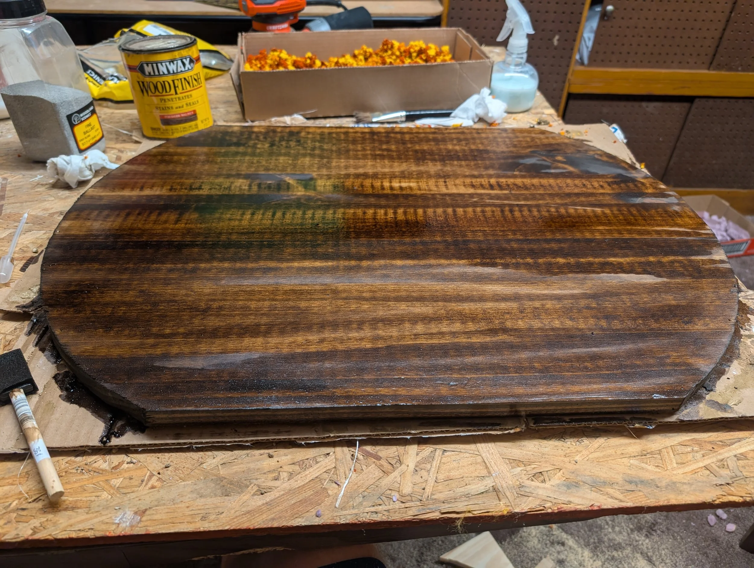 A round wooden tabletop with a stained and sealed finish on a workbench surrounded by woodworking supplies, including a can of Minwax wood finish, sandpaper, and a spray bottle.