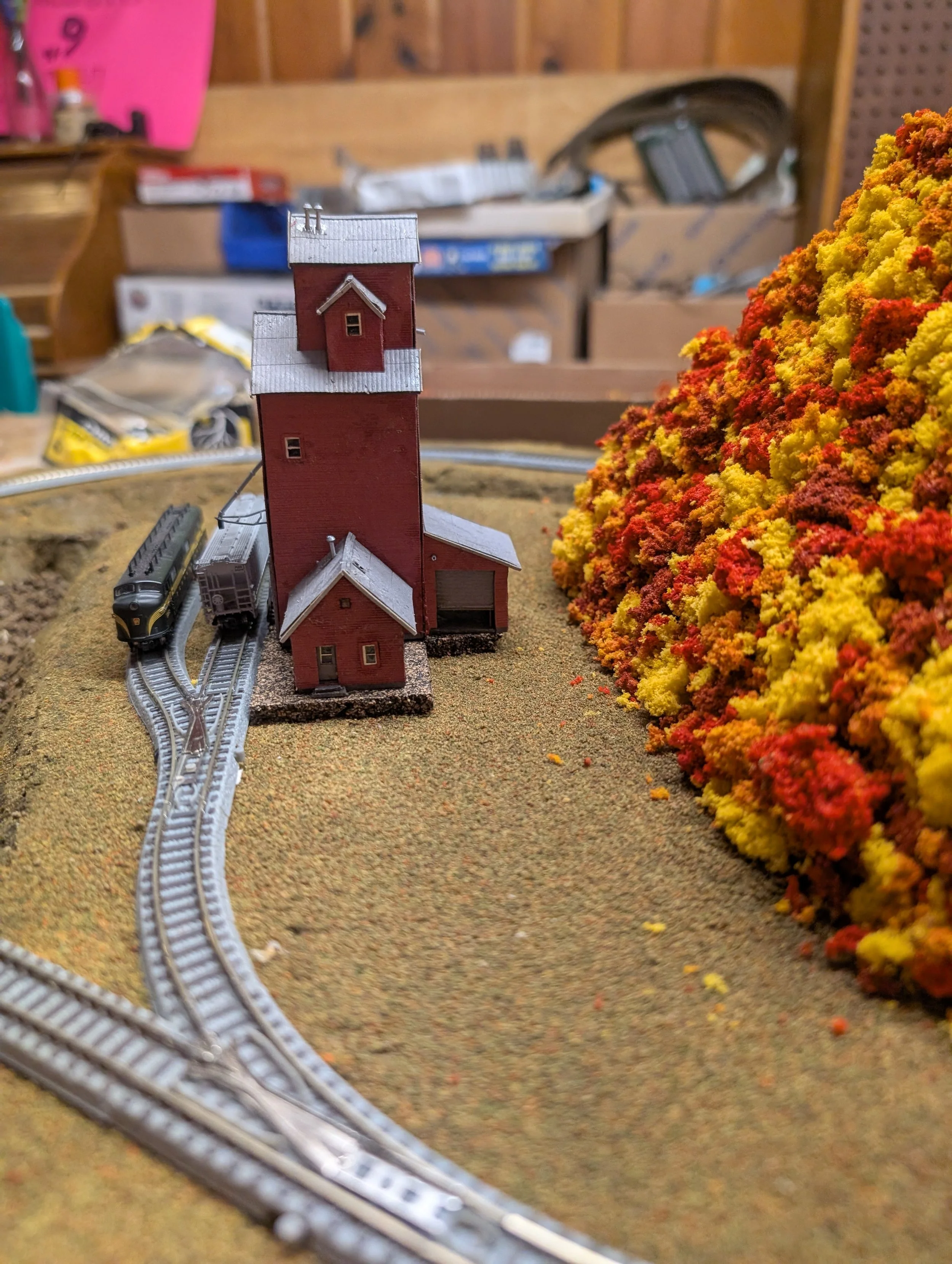 Model train set with a red building, tracks, and a colorful autumn tree landscape.