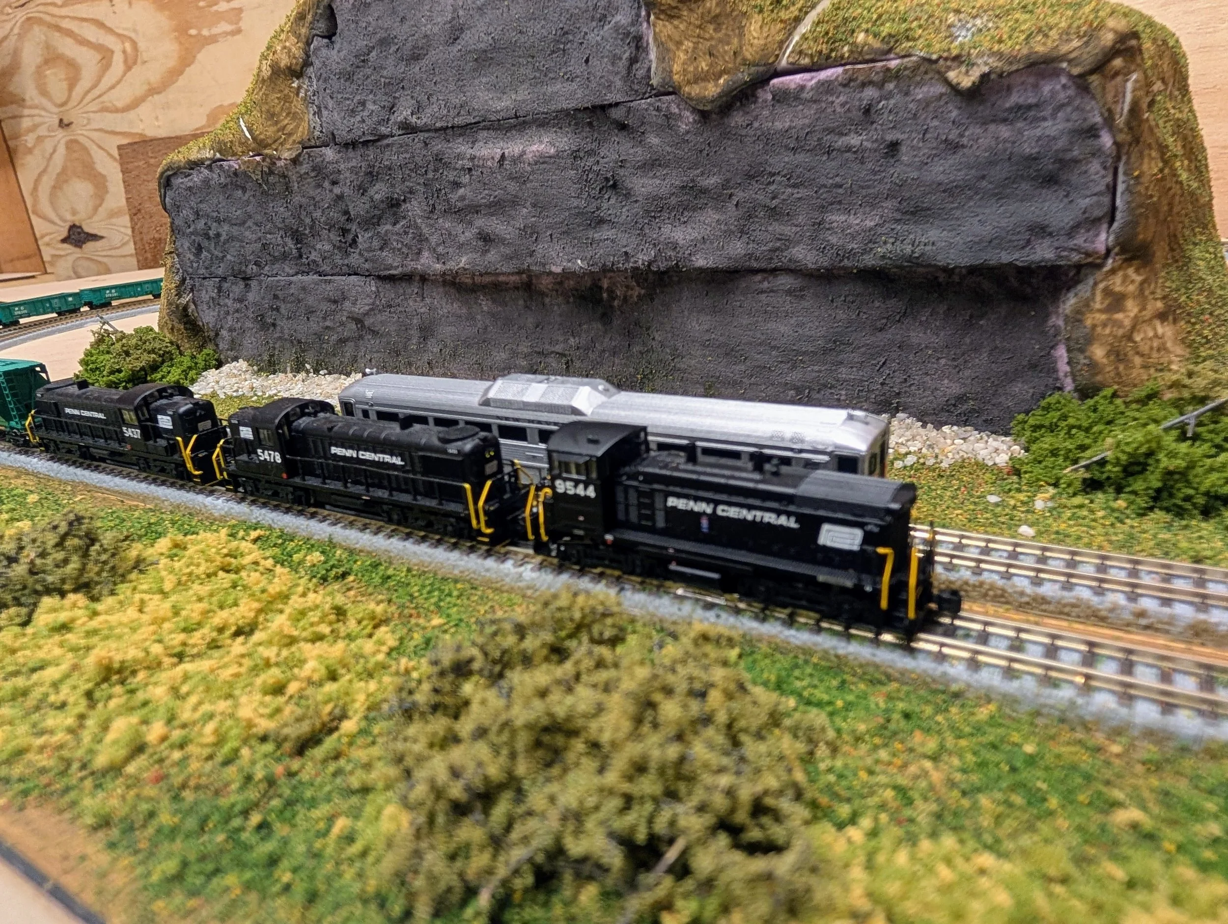Model train set with black locomotives and passenger car on tracks near a rocky hillside with greenery.