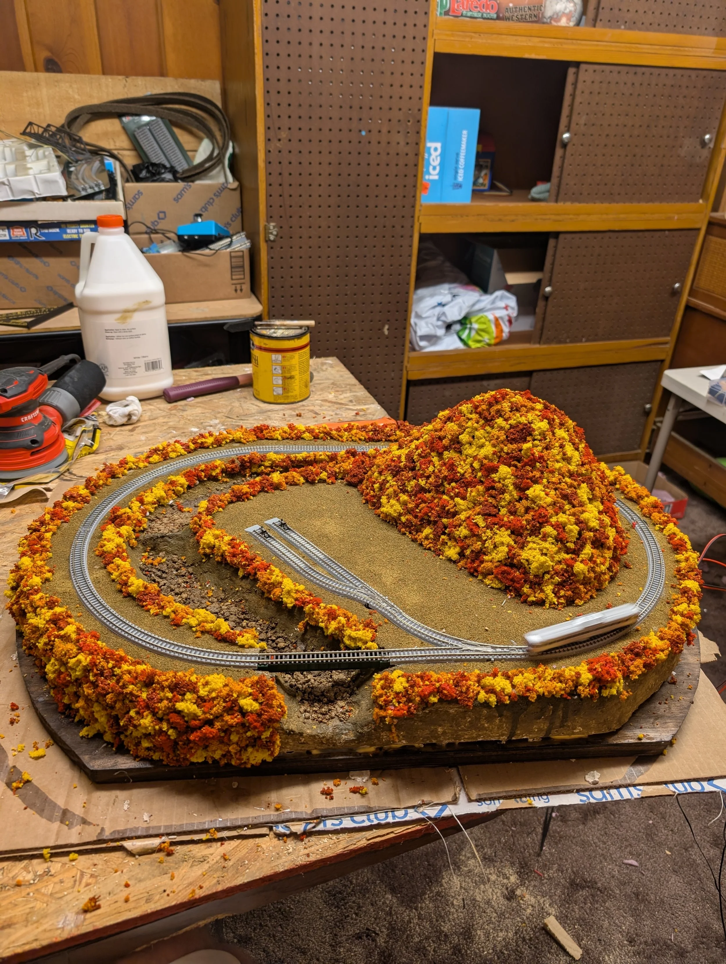 A model train layout featuring a mountain with colorful foliage, train tracks, and a train station, set on a wooden table in a workshop or hobby room.