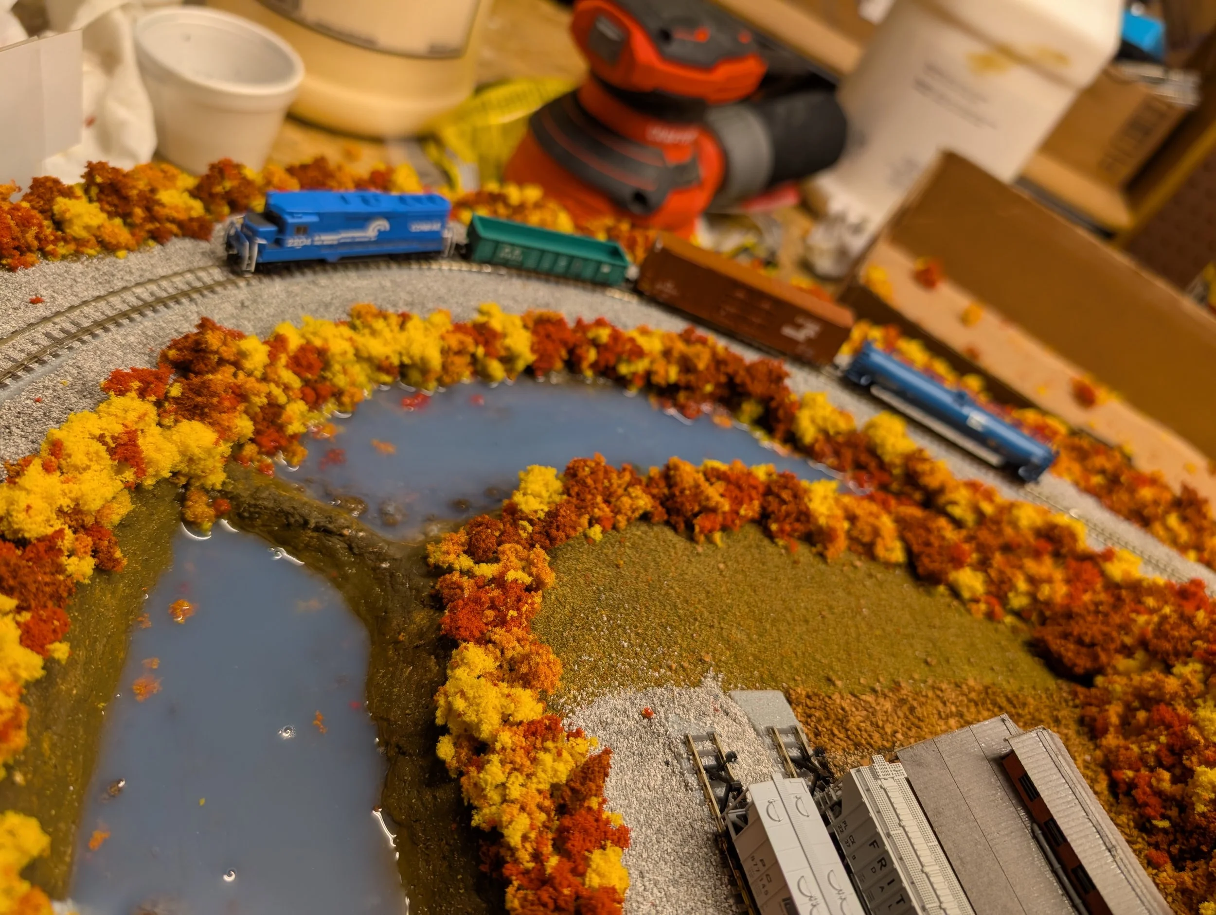 A model train set with a blue locomotive, brown freight cars, and a curved track surrounded by orange, yellow, and red foliage, with a small pond and model buildings.