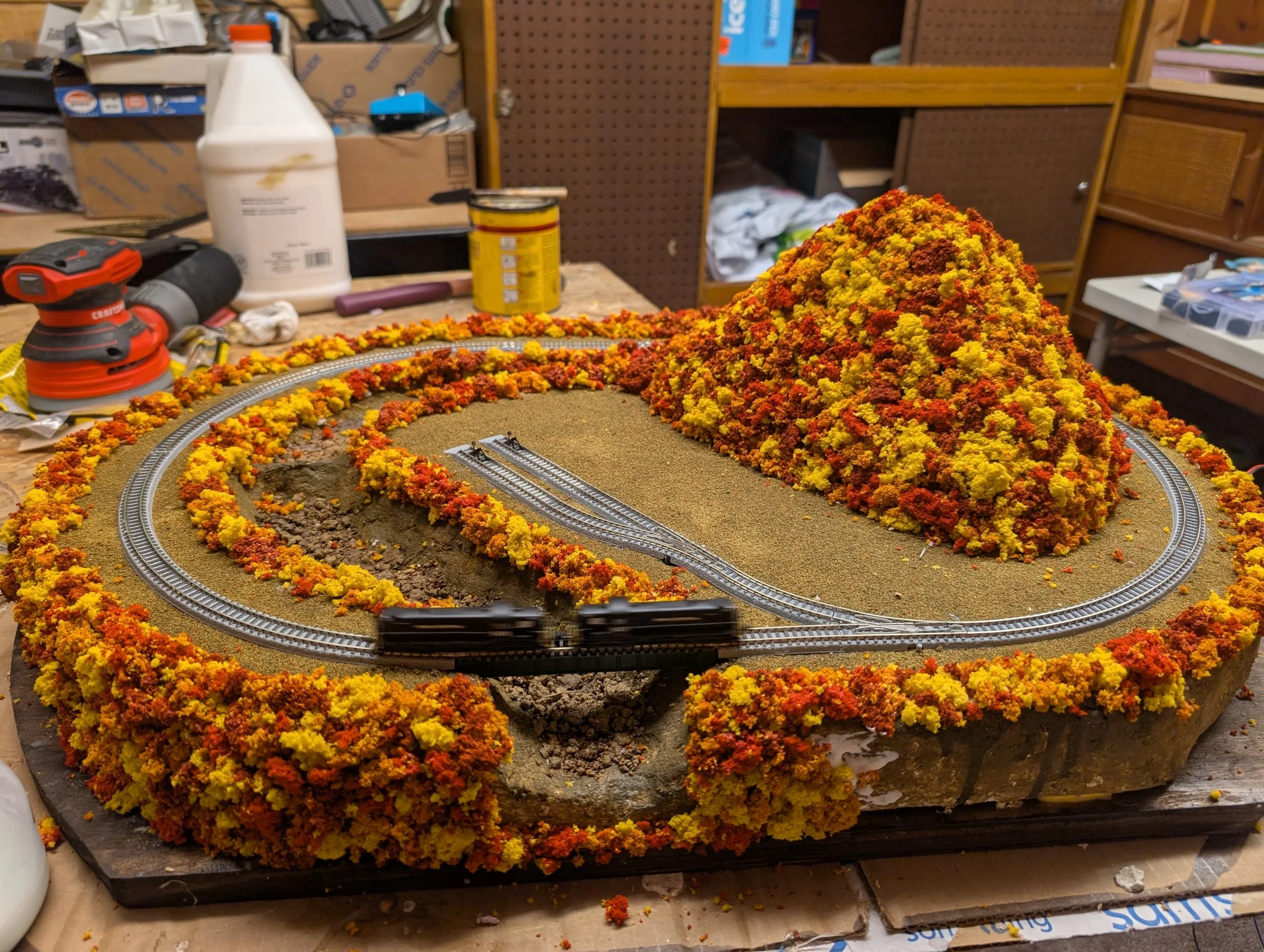 Model train layout with a mountain covered in yellow and orange foliage, track loops around the landscape, on a worktable with tools and supplies in the background.