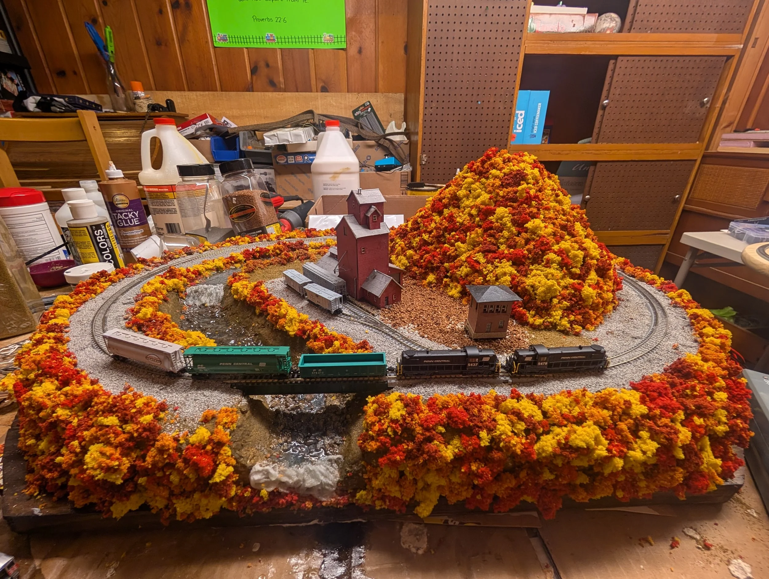 Model train set on a table with a mountain of yellow, orange, and red foliage, a pond, a red barn, and train tracks. The table is in a workshop or hobby room with shelves and various supplies in the background.