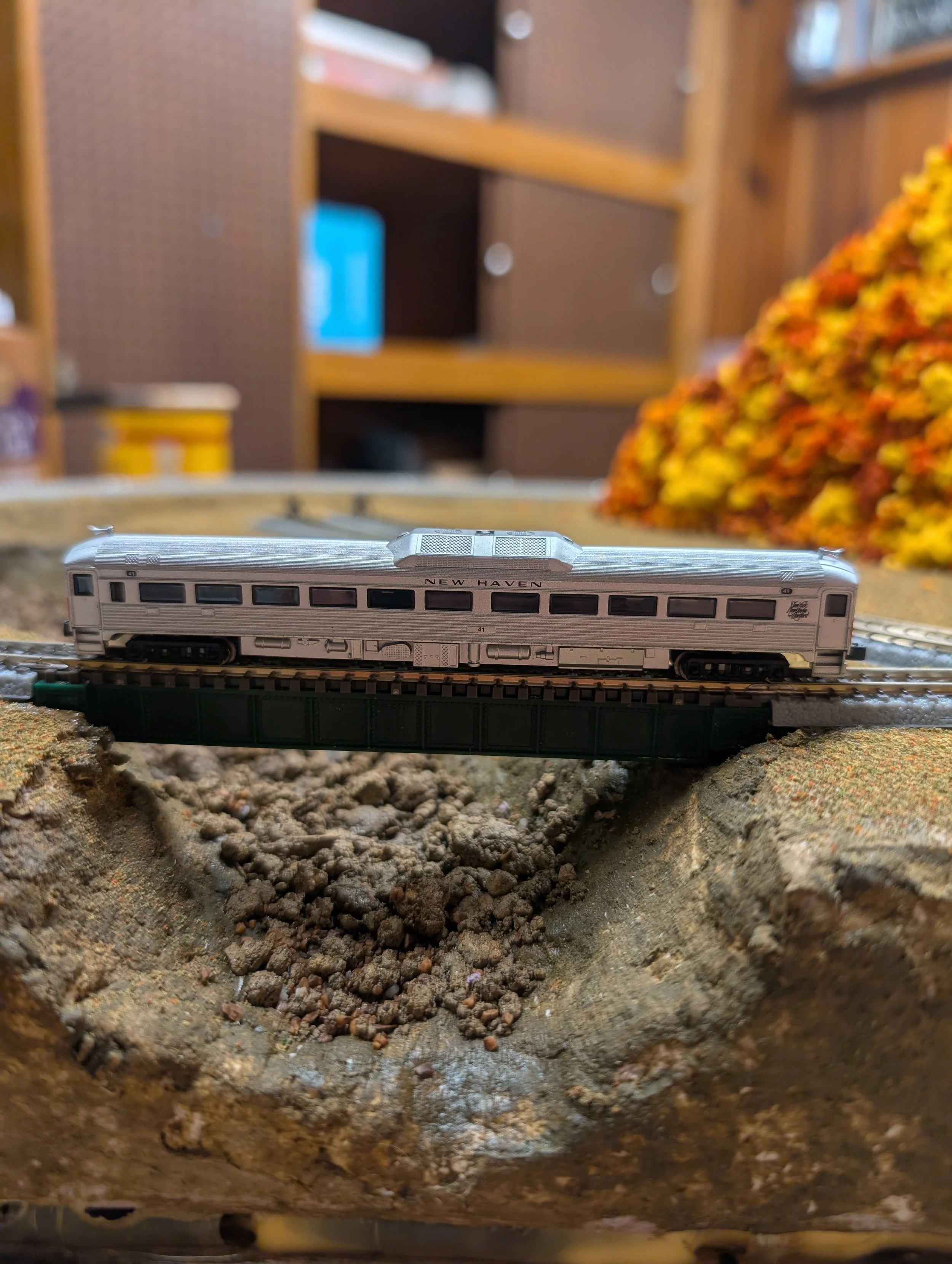 A detailed model train car labeled "NEW HAVEN" on a miniature railroad track, set on a terrain with rocks and dirt, with a blurred background of wooden shelves and a colorful autumn-themed decoration.