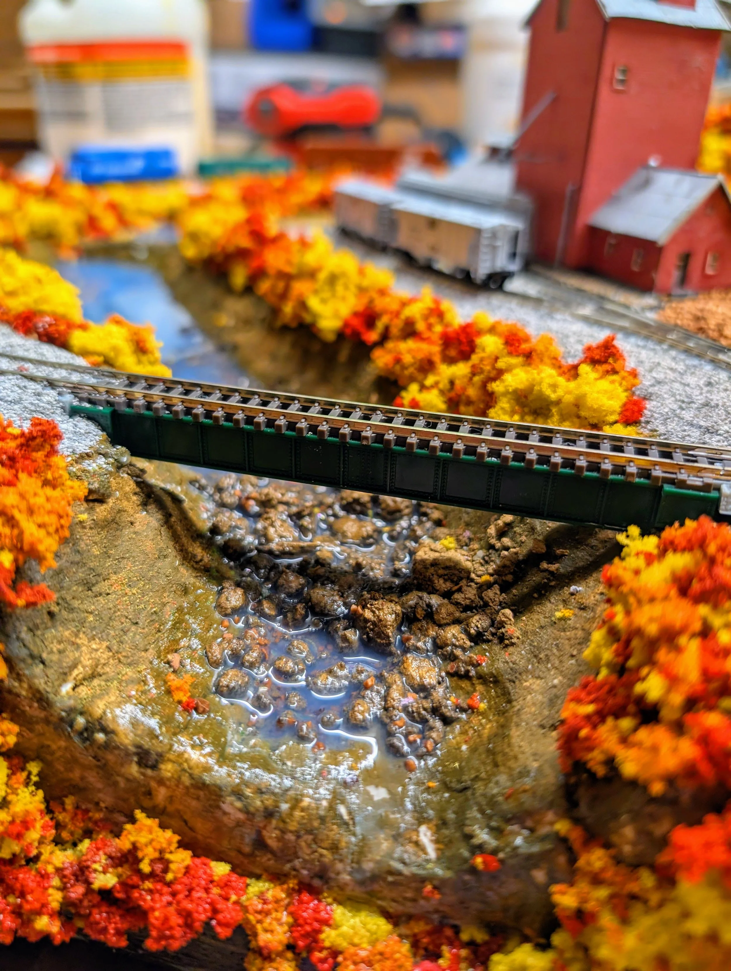 Detailed model of a rural train station scene with a bridge over a creek, colorful autumn trees, a red barn, and a train car in the background.
