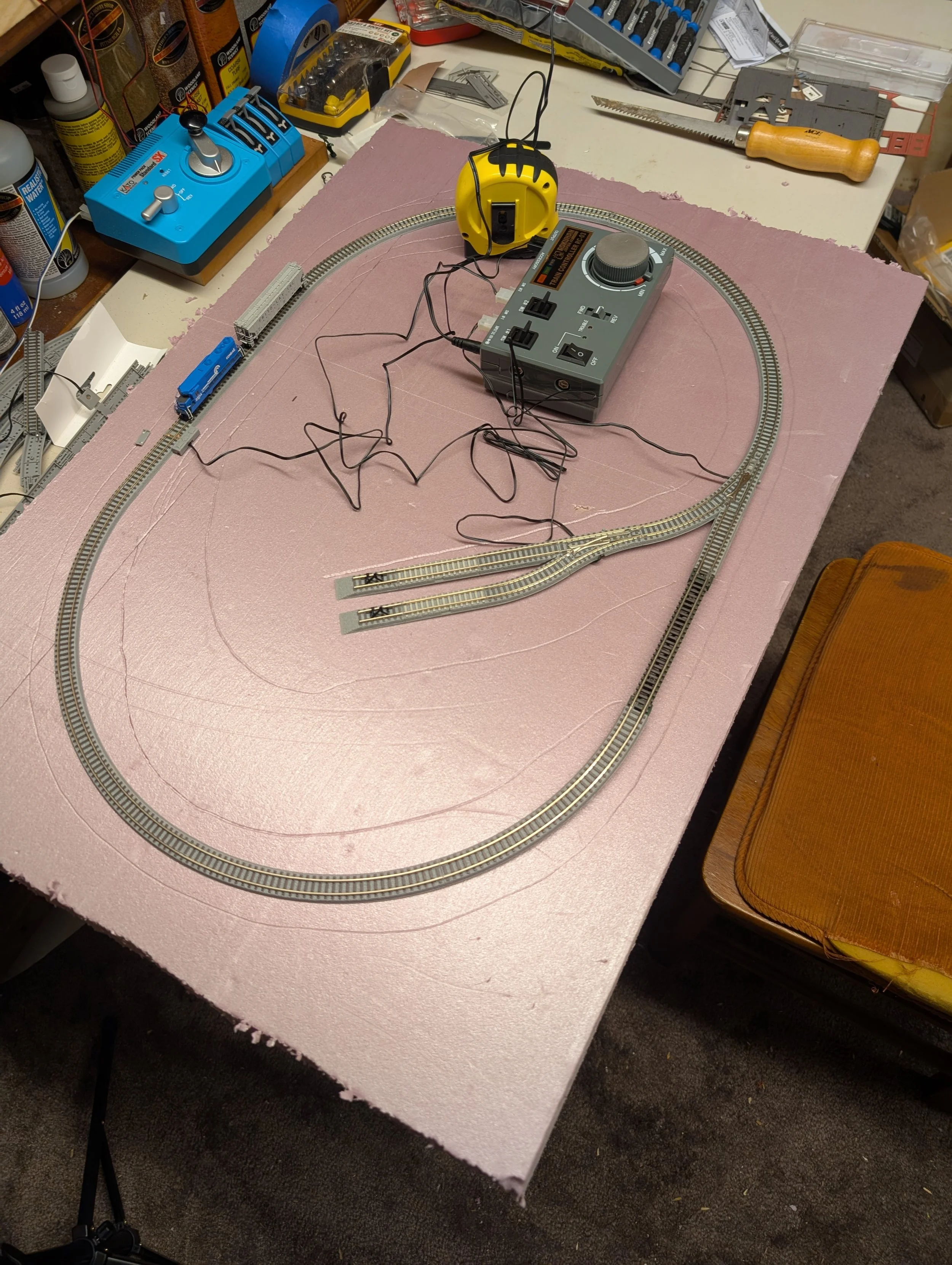A model train setup on a pink foam base, with tracks forming an oval and a switch track section. There is a control box with wires connected to the tracks, a yellow tape measure, various tools, and supplies on the workbench around the setup.