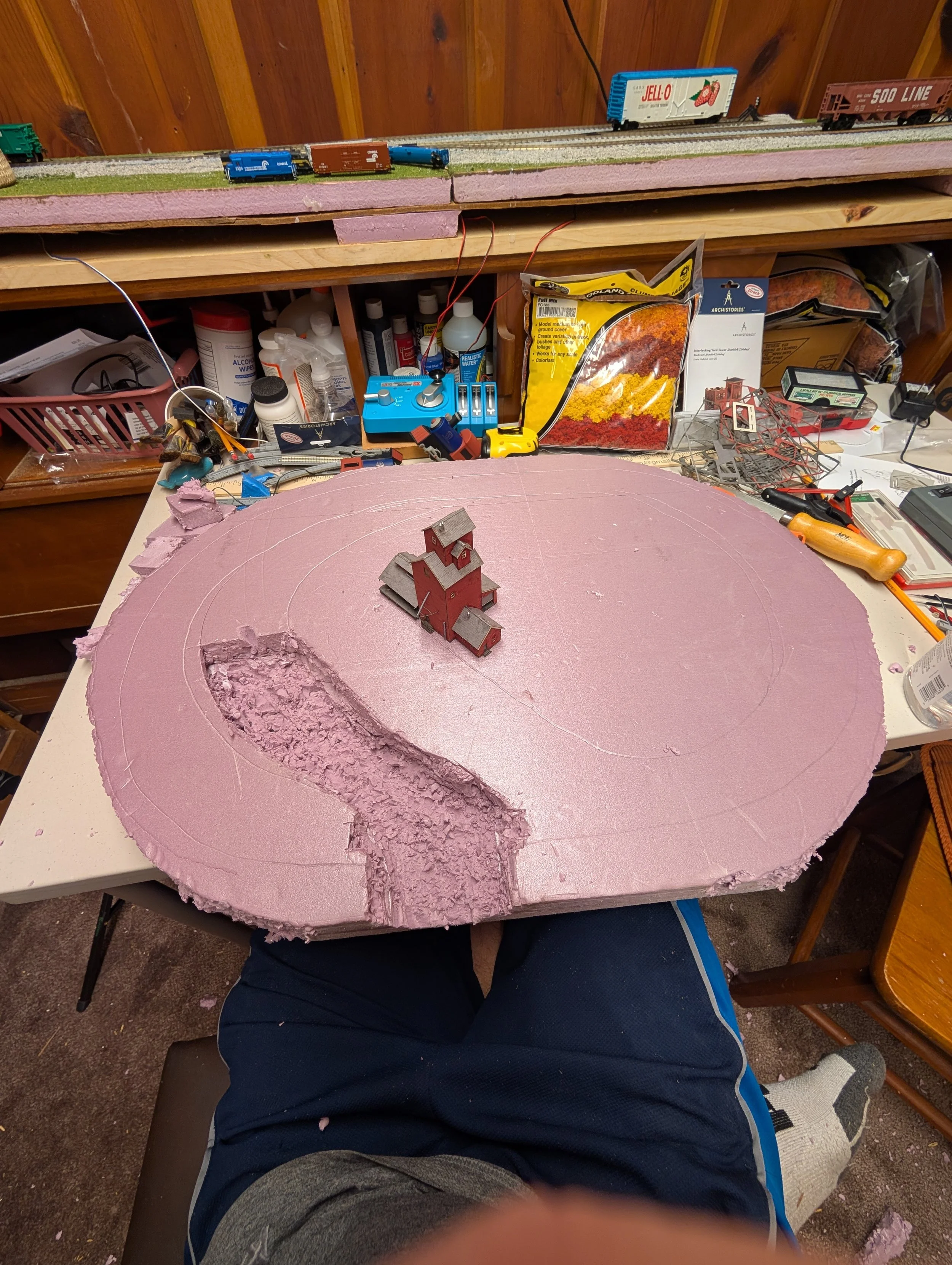 DIY model train layout under construction on a work table with pink foam base, a small red barn, and various tools and supplies surrounding it.