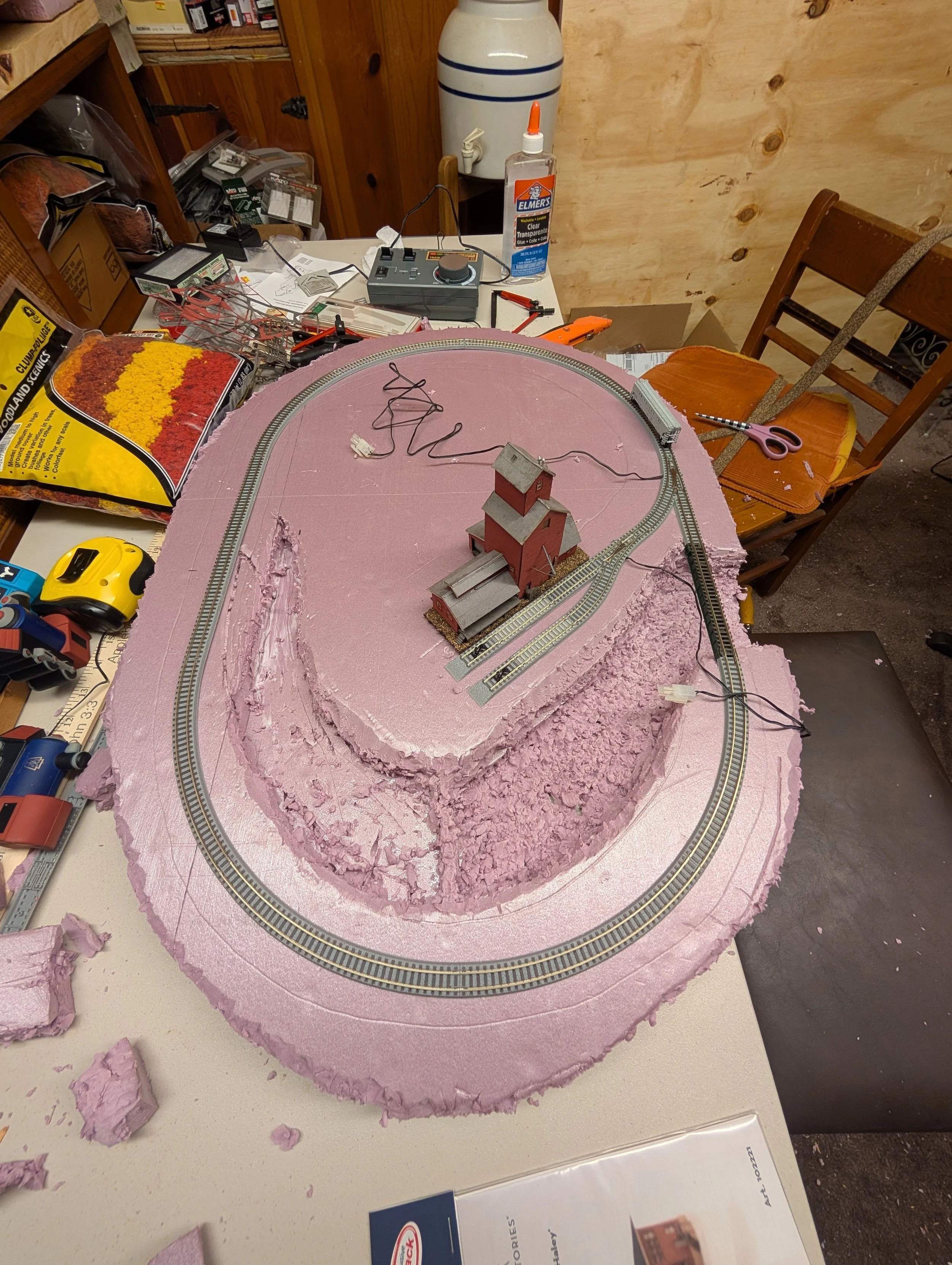 A model train layout under construction showing a circular track on pink foam terrain with a small two-story red and gray building, wires, tools, and modeling supplies scattered around a cluttered worktable.