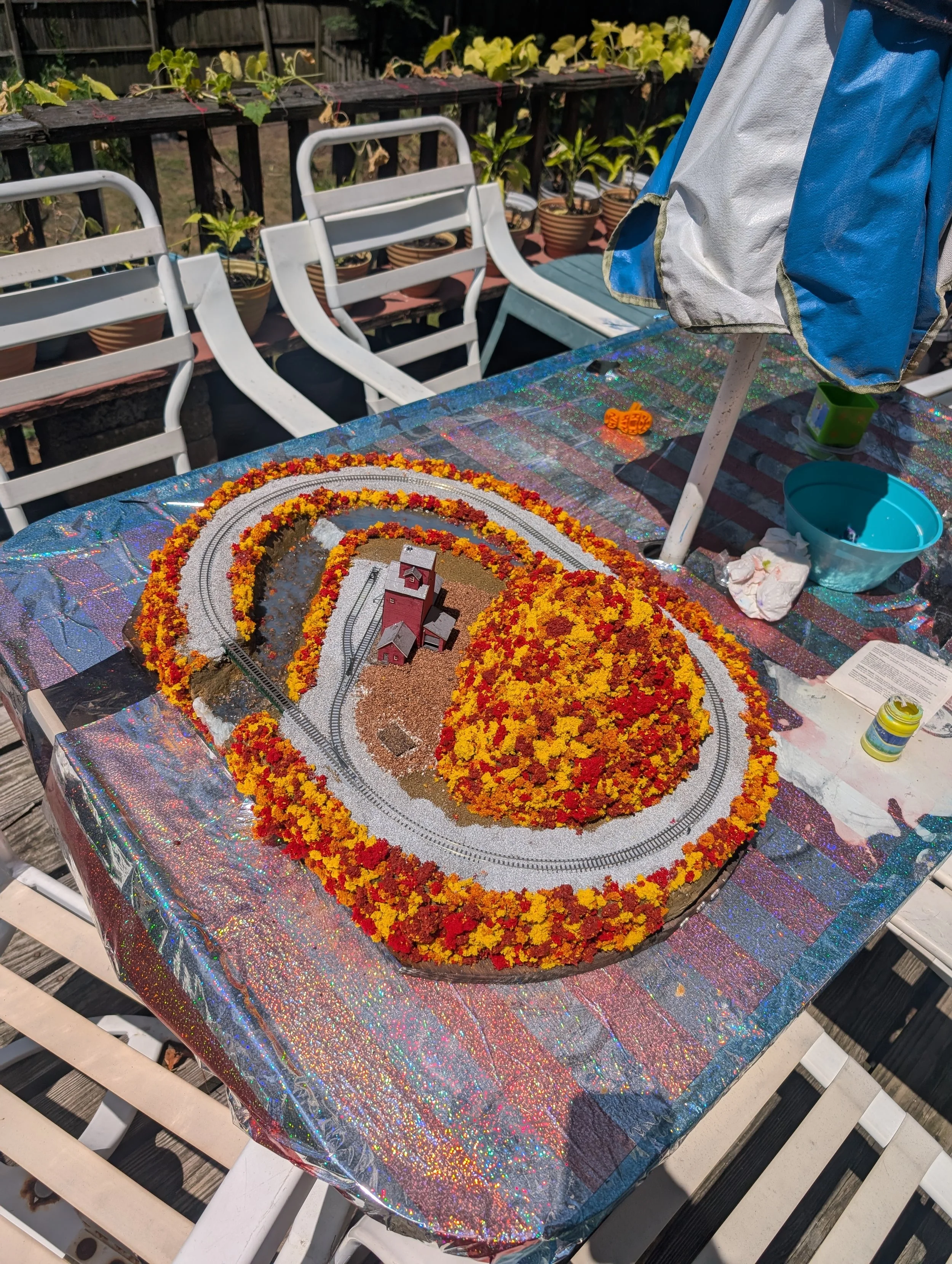 A detailed model train layout decorated with vibrant yellow and red flowers, with houses and tracks, set on a table outdoors.