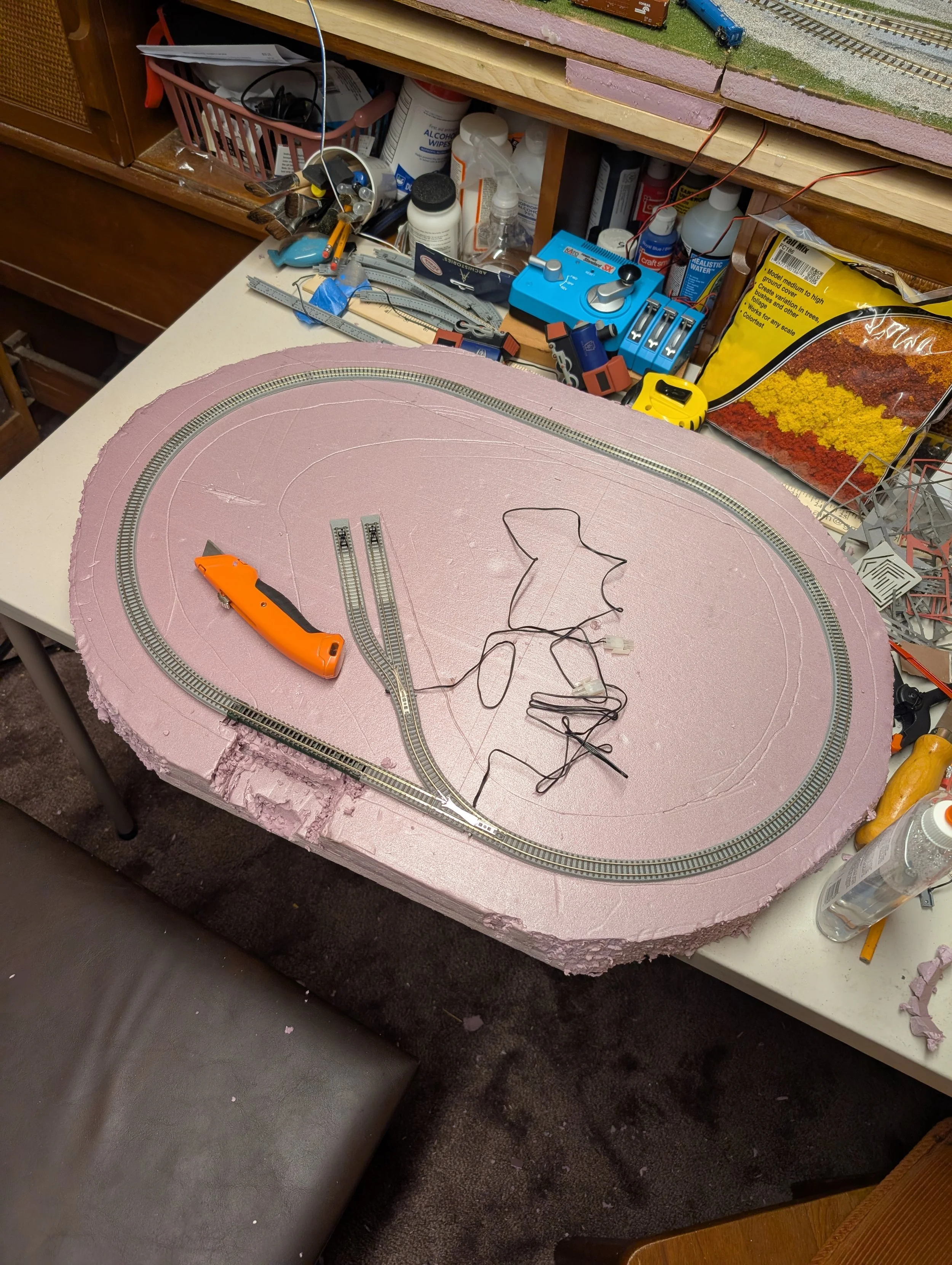 Model train track being assembled on pink foam base with tools and tools and supplies around it.